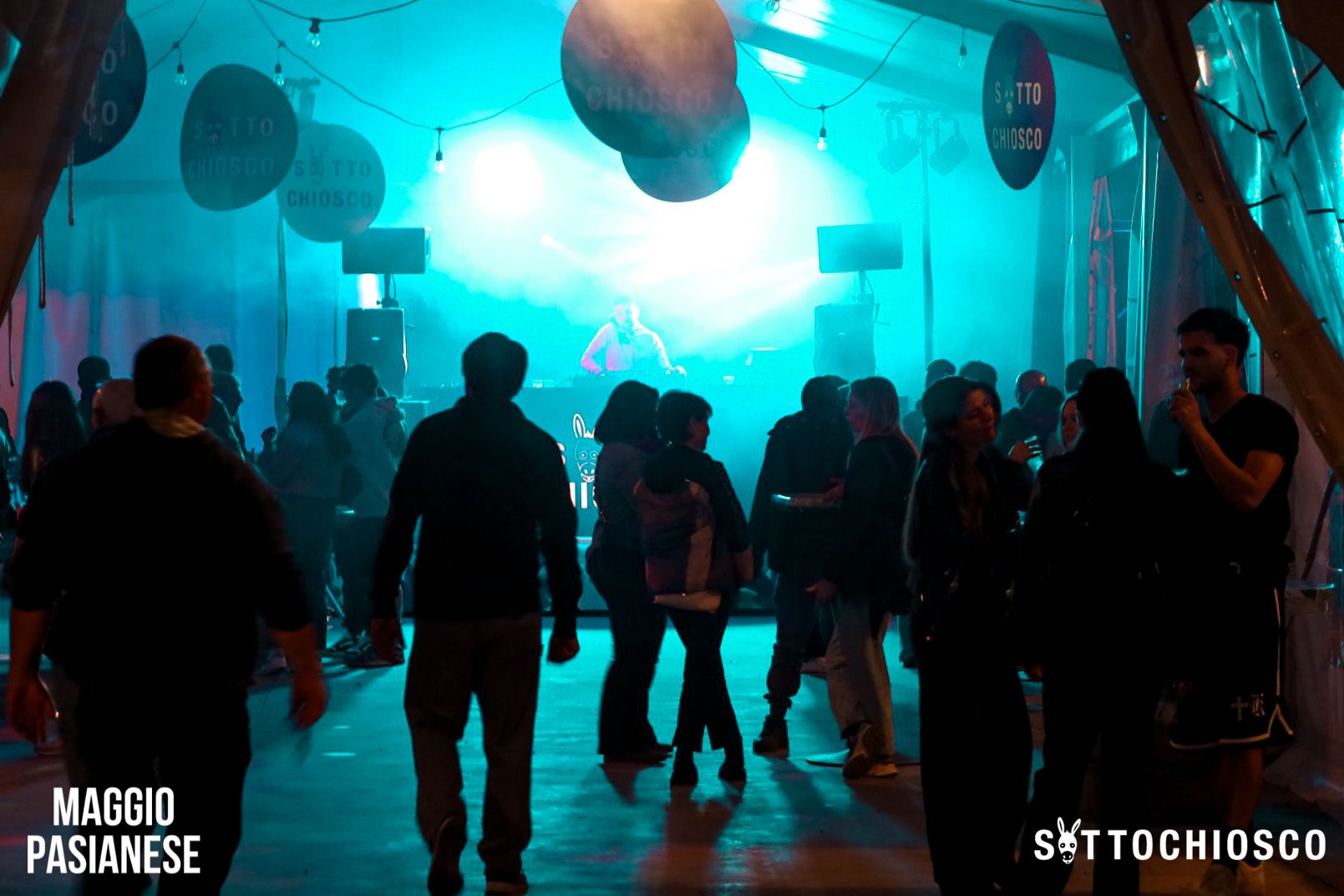 Un gruppo di persone socializza e balla in un locale interno scarsamente illuminato da luci blu, con un DJ sul palco e cartelli rotondi appesi al soffitto. Sono visibili i loghi dell'evento "Maggio Pasianese" e "Sottochiosco".