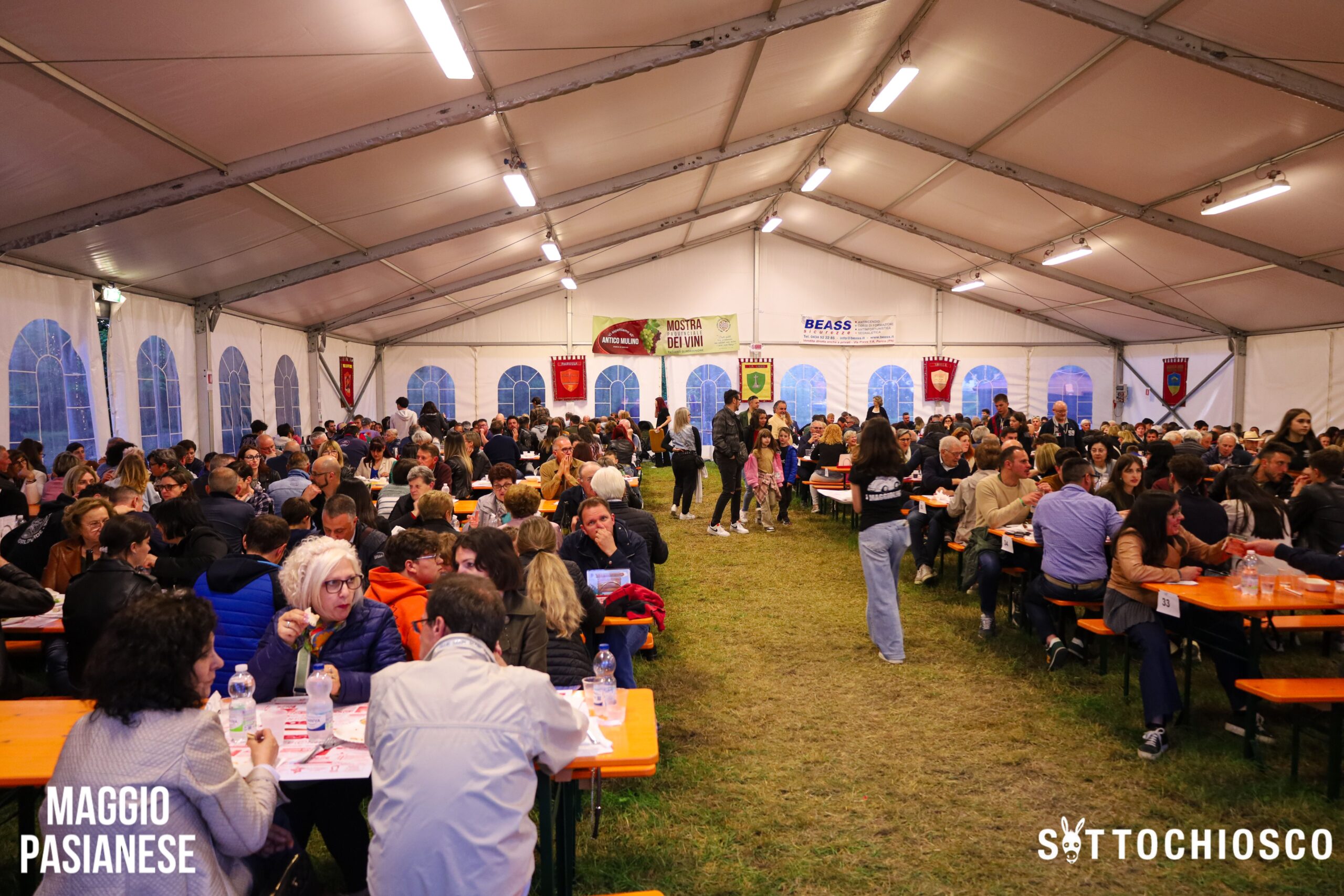Un folto gruppo di persone siede a lunghi tavoli all'interno di una tenda, gustando cibo e chiacchierando durante un vivace evento festivo. Striscioni pendono dal soffitto e l'atmosfera è calda e conviviale.