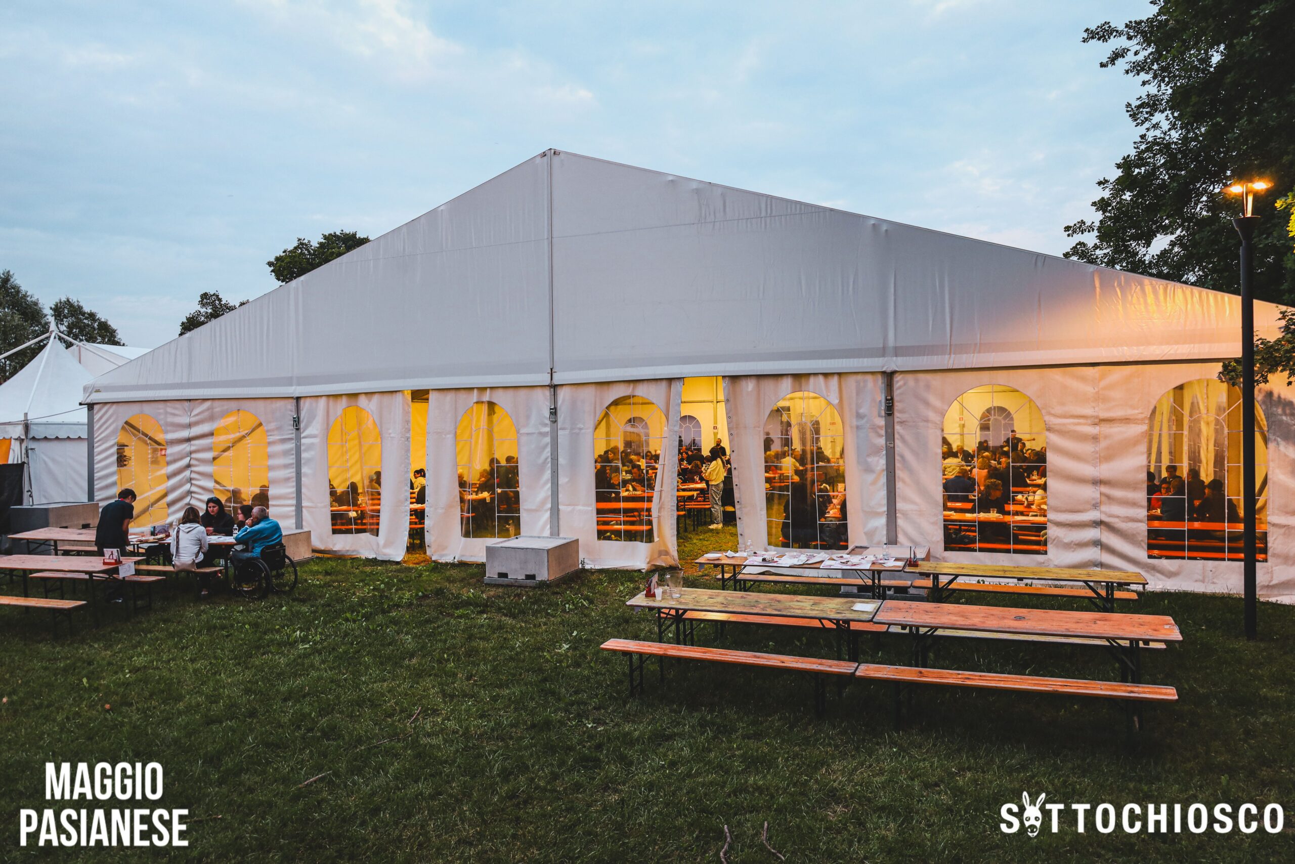 Grande tenda bianca per eventi con finestre ad arco, illuminata da una luce calda all'interno, con persone visibili attraverso le finestre. All'esterno, tavoli da picnic e alcune persone sedute sul prato. Sono visibili i loghi "MAGGIO PASIANESE" e "SOTTOCHIOSCO".