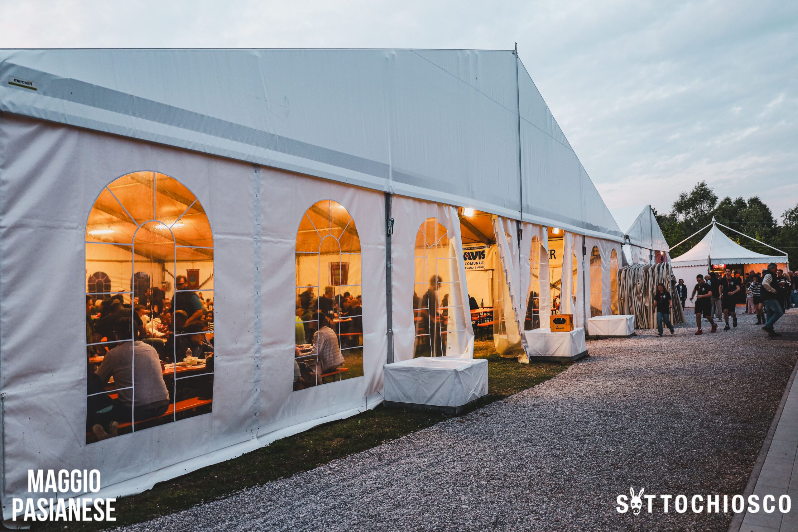 Grande tenda bianca per eventi con finestre ad arco, piena di persone sedute ai tavoli all'interno. La tenda è allestita all'aperto su ghiaia, con persone che camminano vicino all'ingresso. Sono visibili i loghi "MAGGIO PASIANESE" e "SOTTOCHIOSCO".