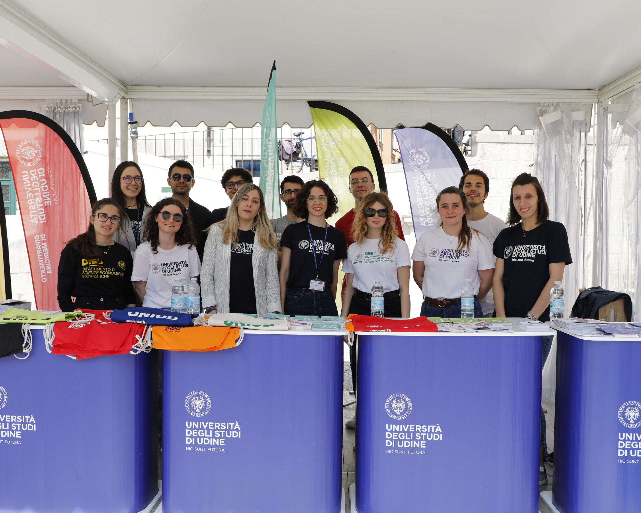 Un gruppo di studenti è in piedi dietro i banchi informativi blu, sotto una tenda bianca, indossando magliette con i loghi dell'università. Sono esposti striscioni colorati e materiale promozionale. Sui banchi si legge "Università degli Studi di Udine".