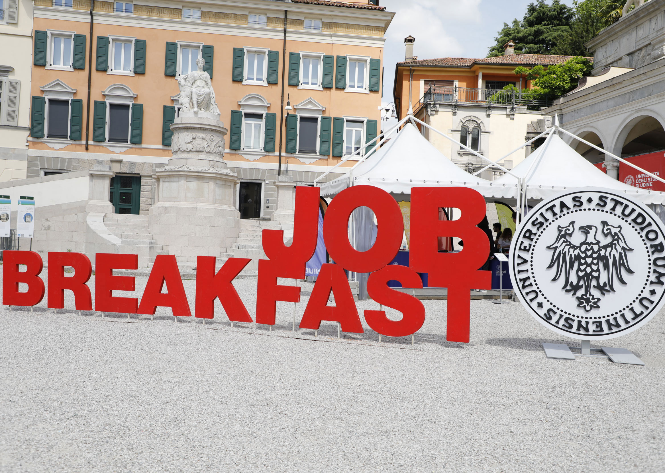 Grandi lettere rosse con la scritta "JOB BREAKFAST" (Colazione di lavoro) sono poste in una piazza all'aperto, vicino allo stemma dell'università, con edifici storici e tende bianche sullo sfondo.