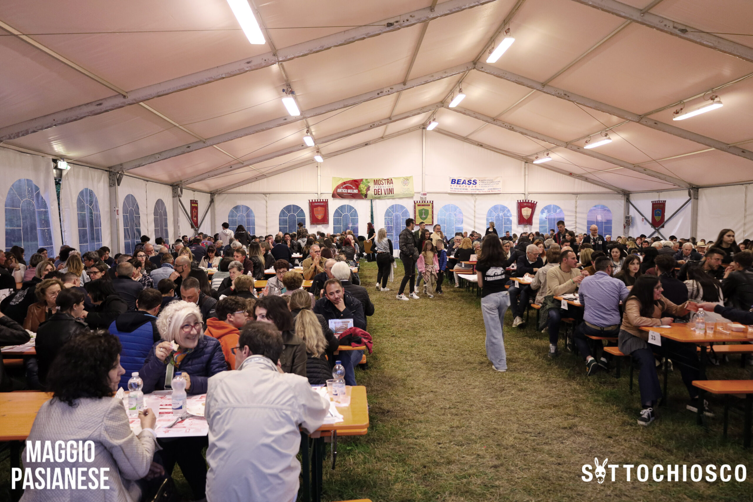 Una grande folla di persone sedute a lunghi tavoli all'interno di una tenda bianca per eventi, che mangiano e socializzano. Striscioni pendono dalle pareti della tenda. Il terreno è erboso. Negli angoli si leggono le scritte "MAGGIO PASIANESE" e "SOTTOCHIOSCO".