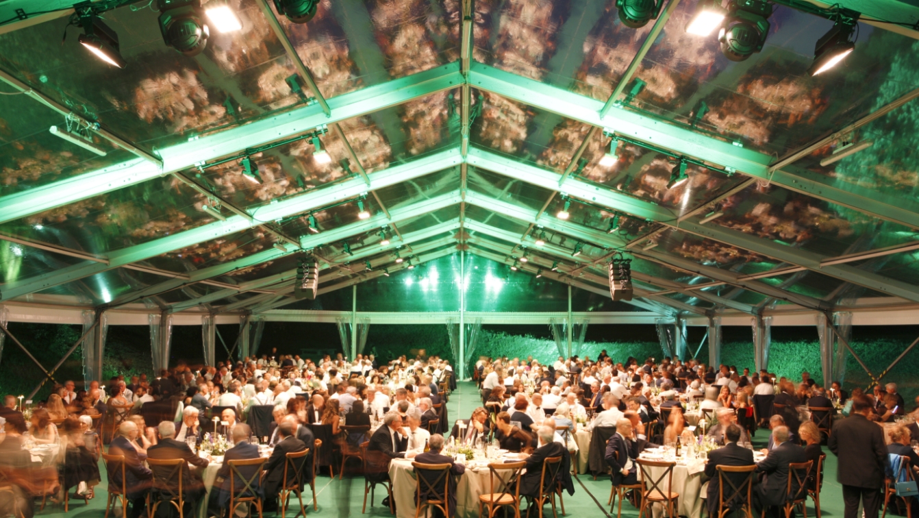 Una grande folla di persone siede ai tavoli rotondi sotto una tenda trasparente illuminata di verde durante un evento formale serale o un banchetto.