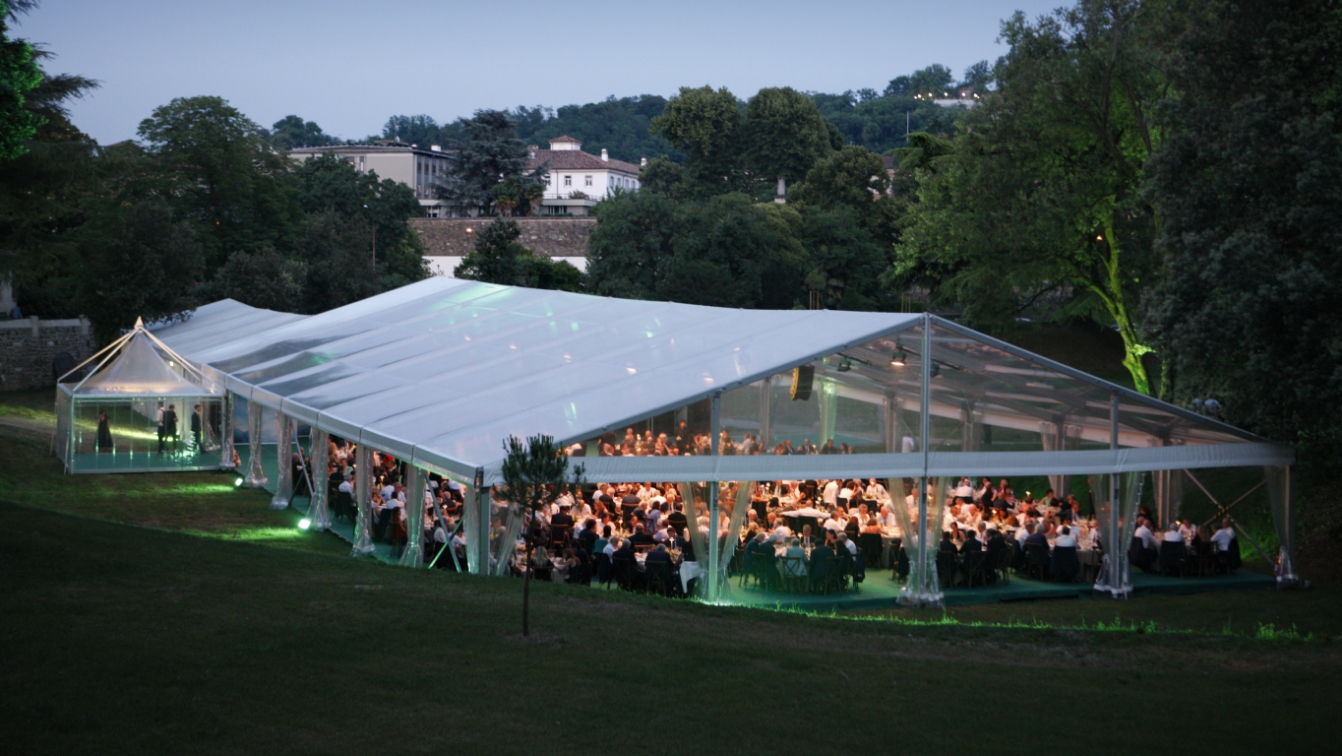 Un grande evento all'aperto si svolge all'imbrunire sotto una tenda trasparente, con molte persone sedute ai tavoli all'interno. La tenda è illuminata da luci soffuse di colore verde e circondata da alberi e piante.