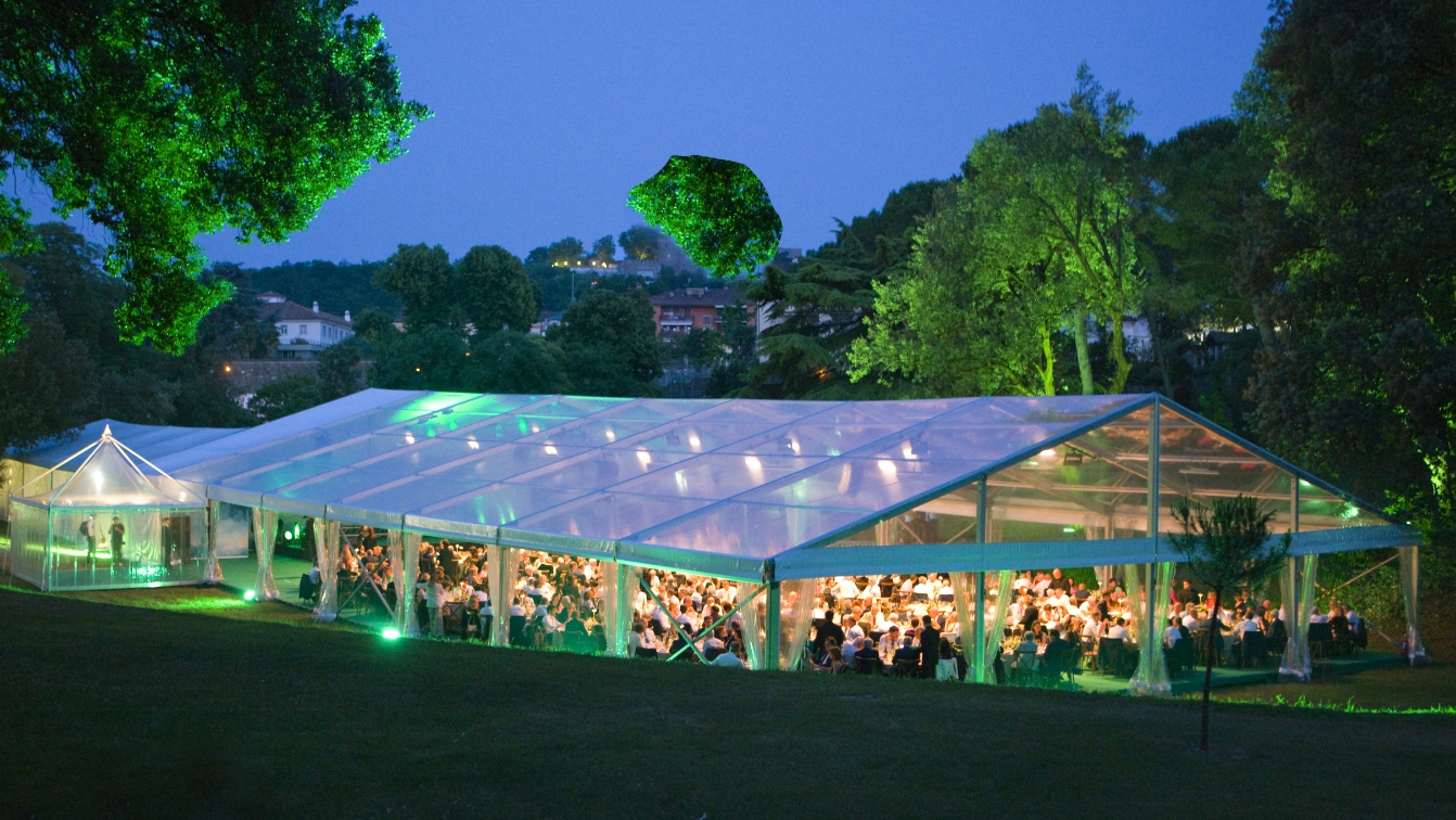 Una grande tenda trasparente allestita all'aperto all'imbrunire, piena di persone sedute ai tavoli sotto una luce calda, circondata da alberi e prati verdi.