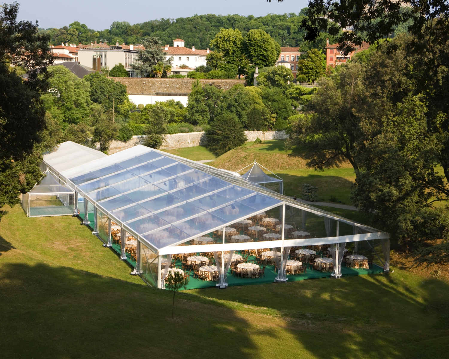 Una grande tenda trasparente allestita su un prato verde con tavoli e sedie rotonde all'interno, circondata da alberi e verde, con edifici e colline sullo sfondo.