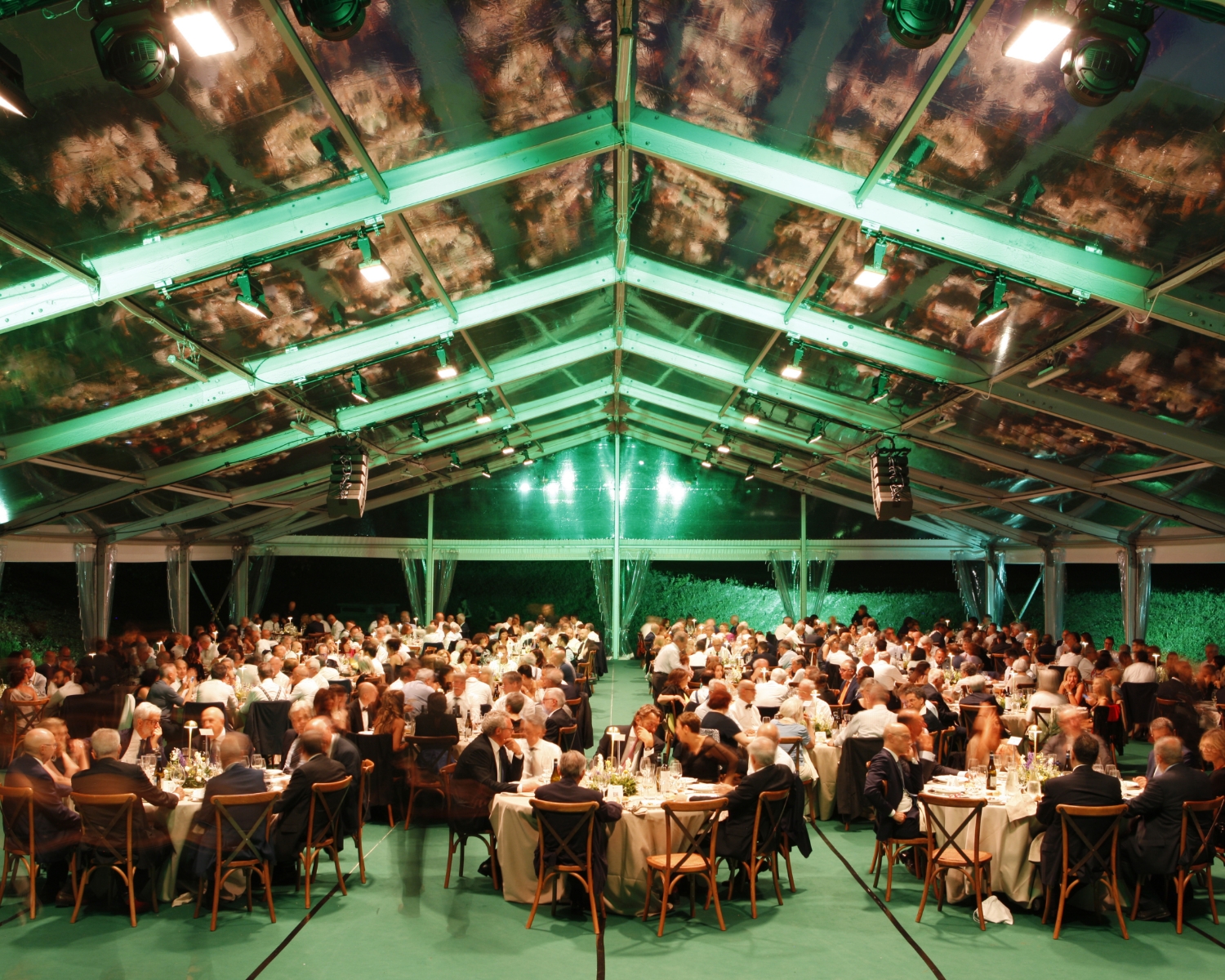Un grande gruppo di persone sedute a tavoli rotondi sotto una tenda trasparente e illuminata di verde in occasione di un evento formale serale o di un banchetto.