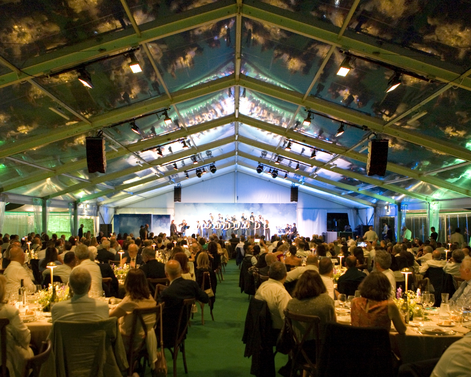 Una grande folla siede a tavoli rotondi sotto una tenda trasparente, guardando una band che si esibisce sul palco. L'evento è formale, con tavoli eleganti, candele e illuminazione ambientale.