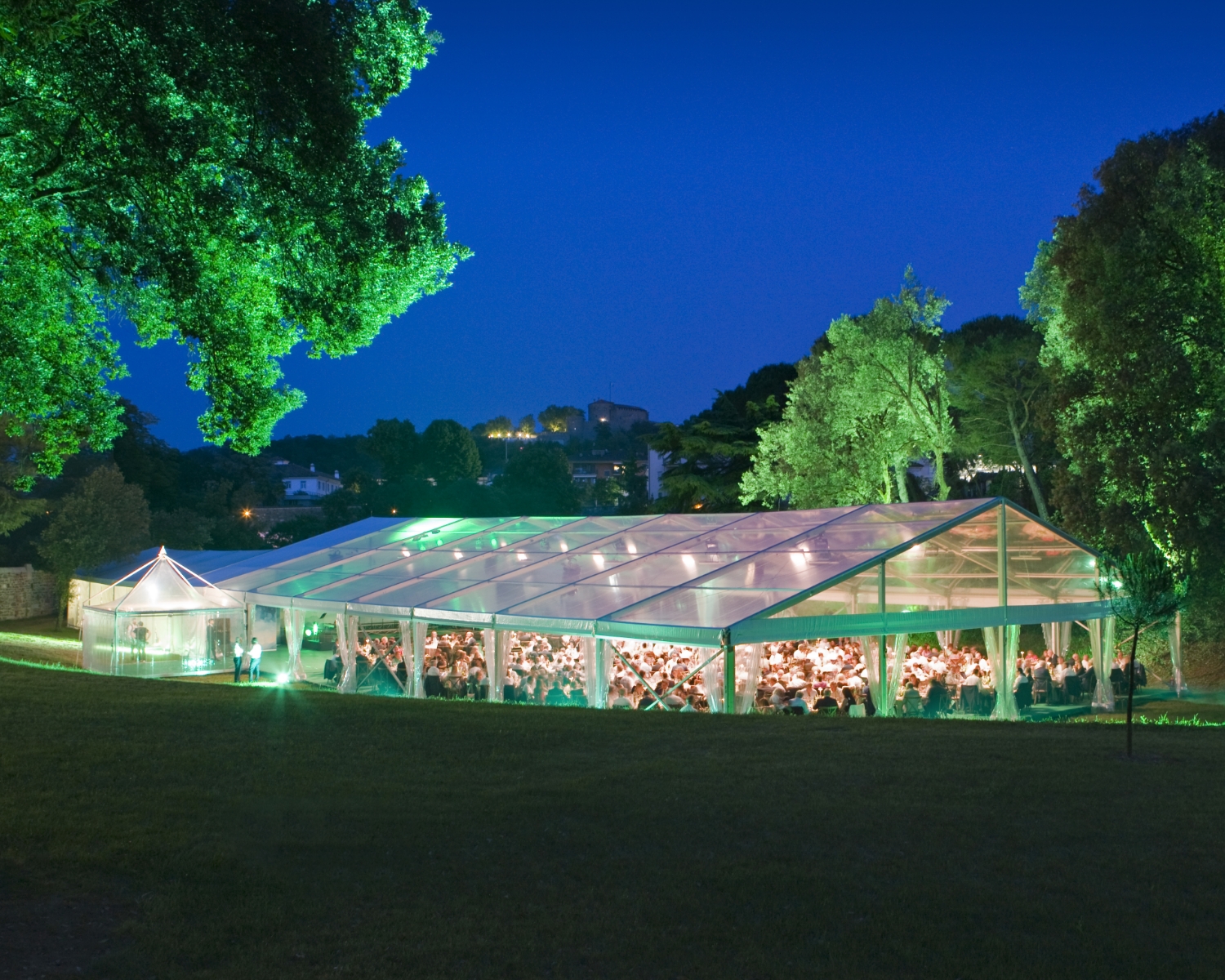 Una grande tenda trasparente si illumina in un'area erbosa all'aperto al tramonto, con persone sedute all'interno. Gli alberi circondano la scena, con il cielo blu intenso e gli edifici lontani visibili sullo sfondo.