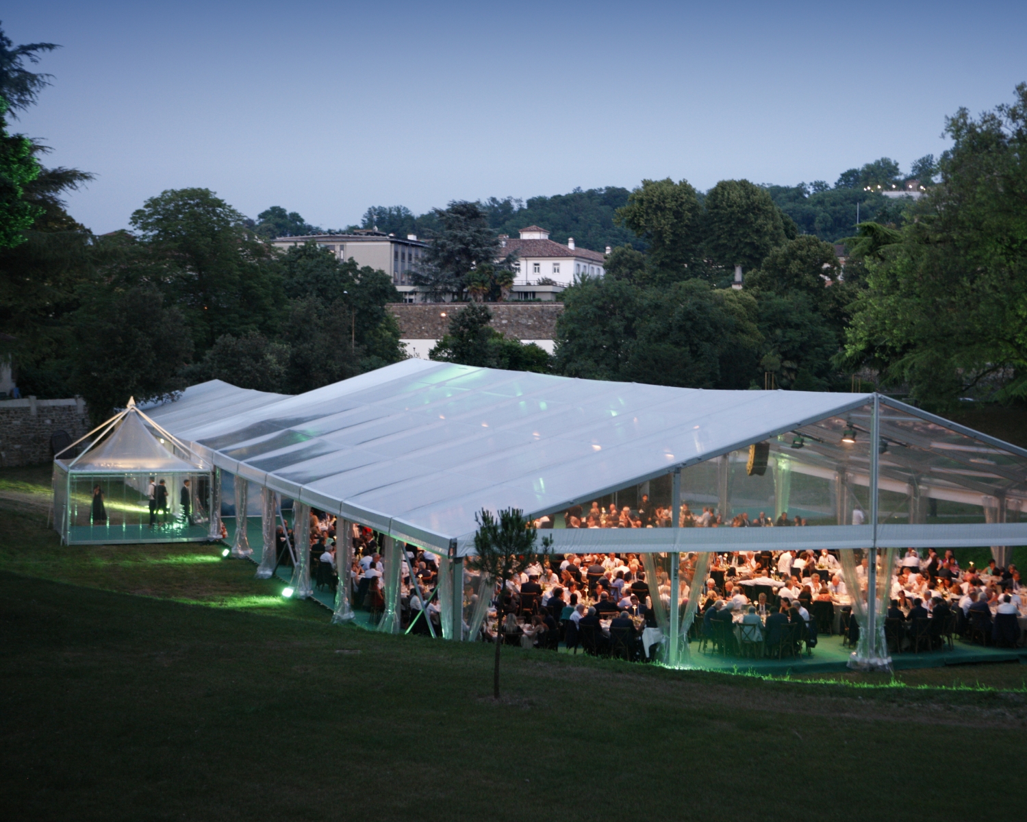 Una grande tenda trasparente allestita all'aperto all'imbrunire, piena di persone sedute a tavoli rotondi per un evento formale, circondata da alberi e colline erbose.
