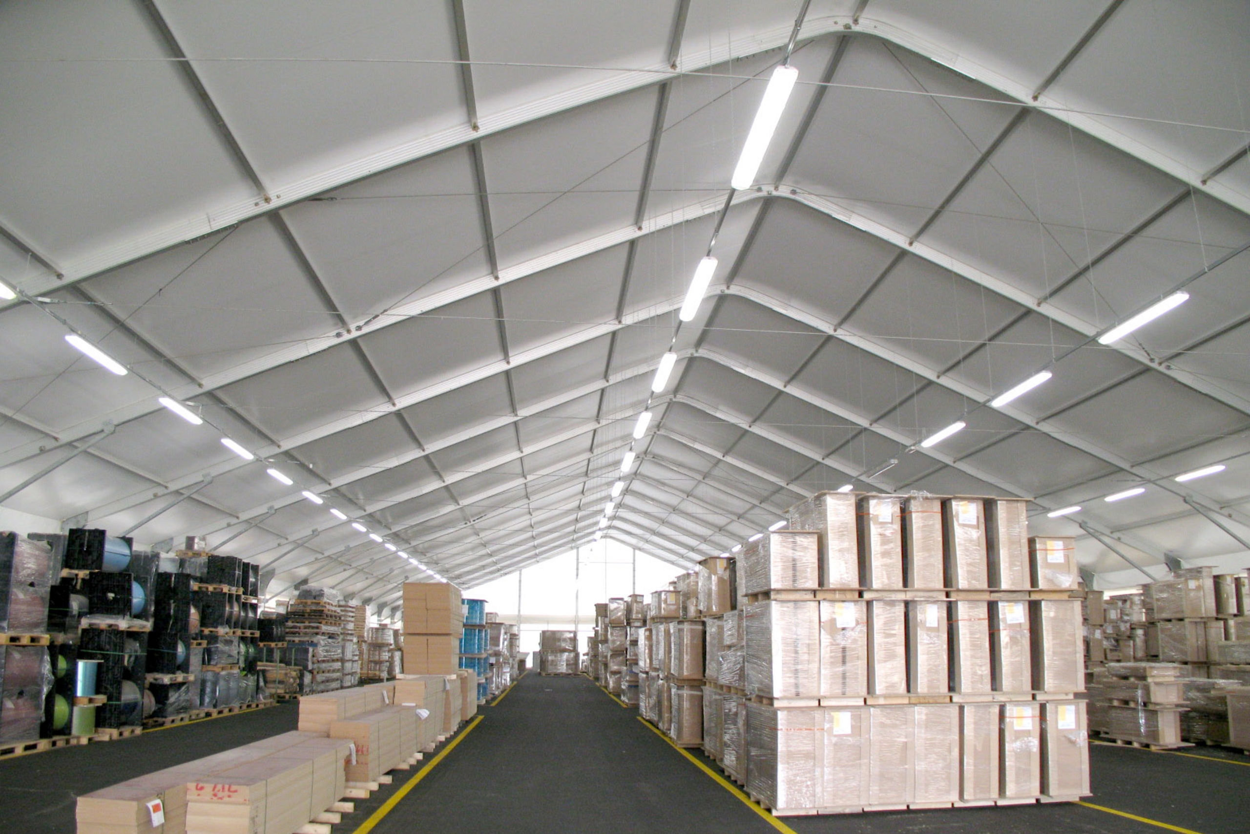Interno di un grande magazzino con alto soffitto bianco, luci fluorescenti e pile di scatole e pallet organizzati in file lungo ampi corridoi. Lo spazio appare luminoso, pulito e ben organizzato.