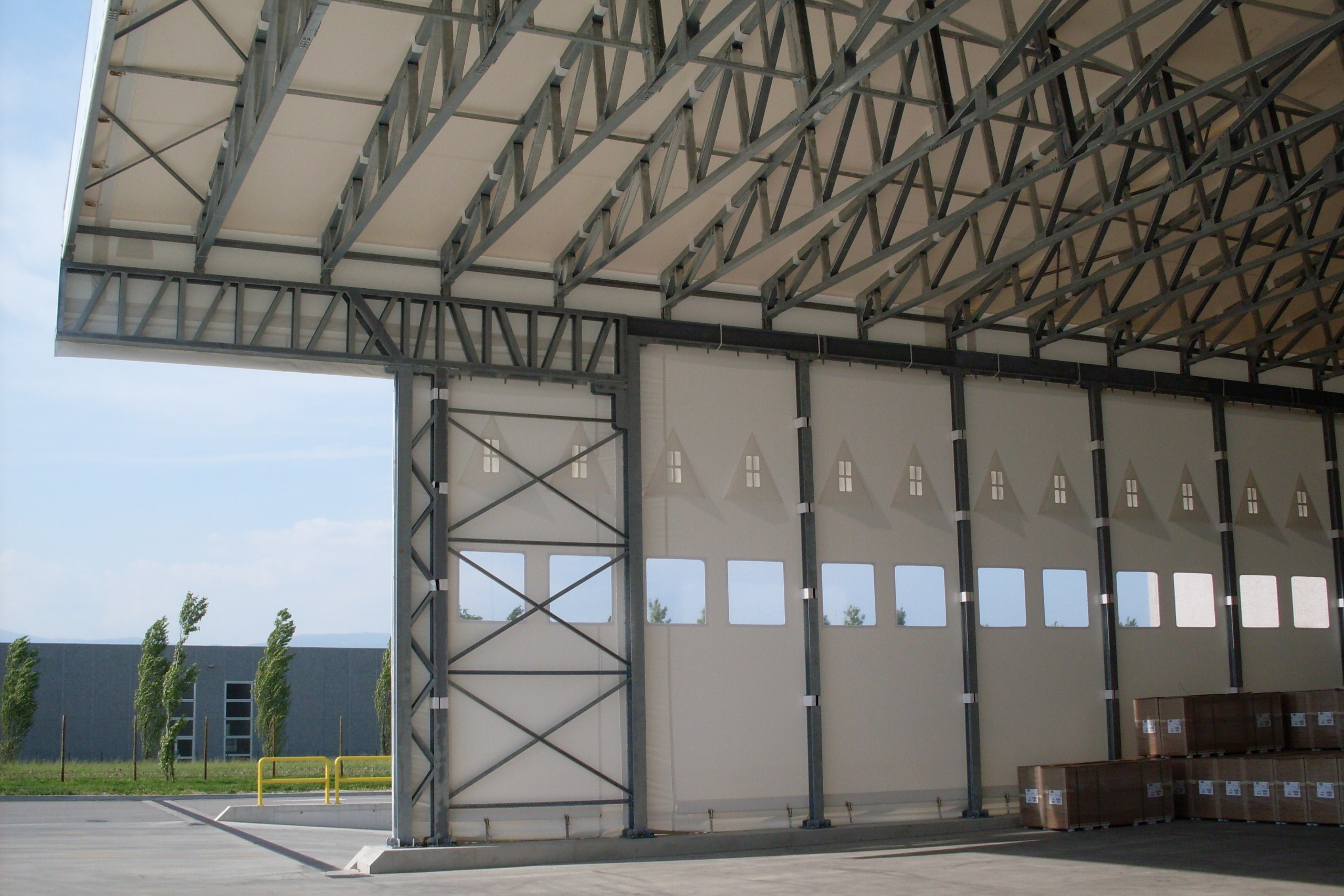 Grande magazzino industriale con porta avvolgibile aperta e parzialmente sollevata, struttura metallica e scatole impilate. L'esterno mostra un cielo sereno, alberi e un edificio grigio sullo sfondo.