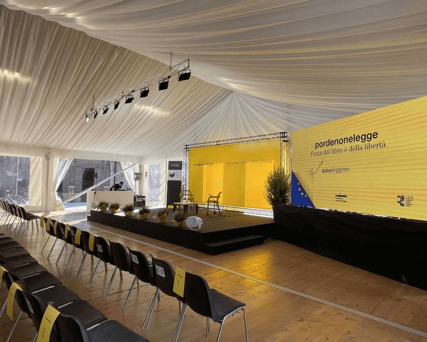 Una grande tenda bianca copre un palcoscenico con fondali gialli con la scritta "pordenonelegge" e file di sedie nere vuote allestite per un evento. L'illuminazione del palco e le piante in vaso decorano lo spazio.