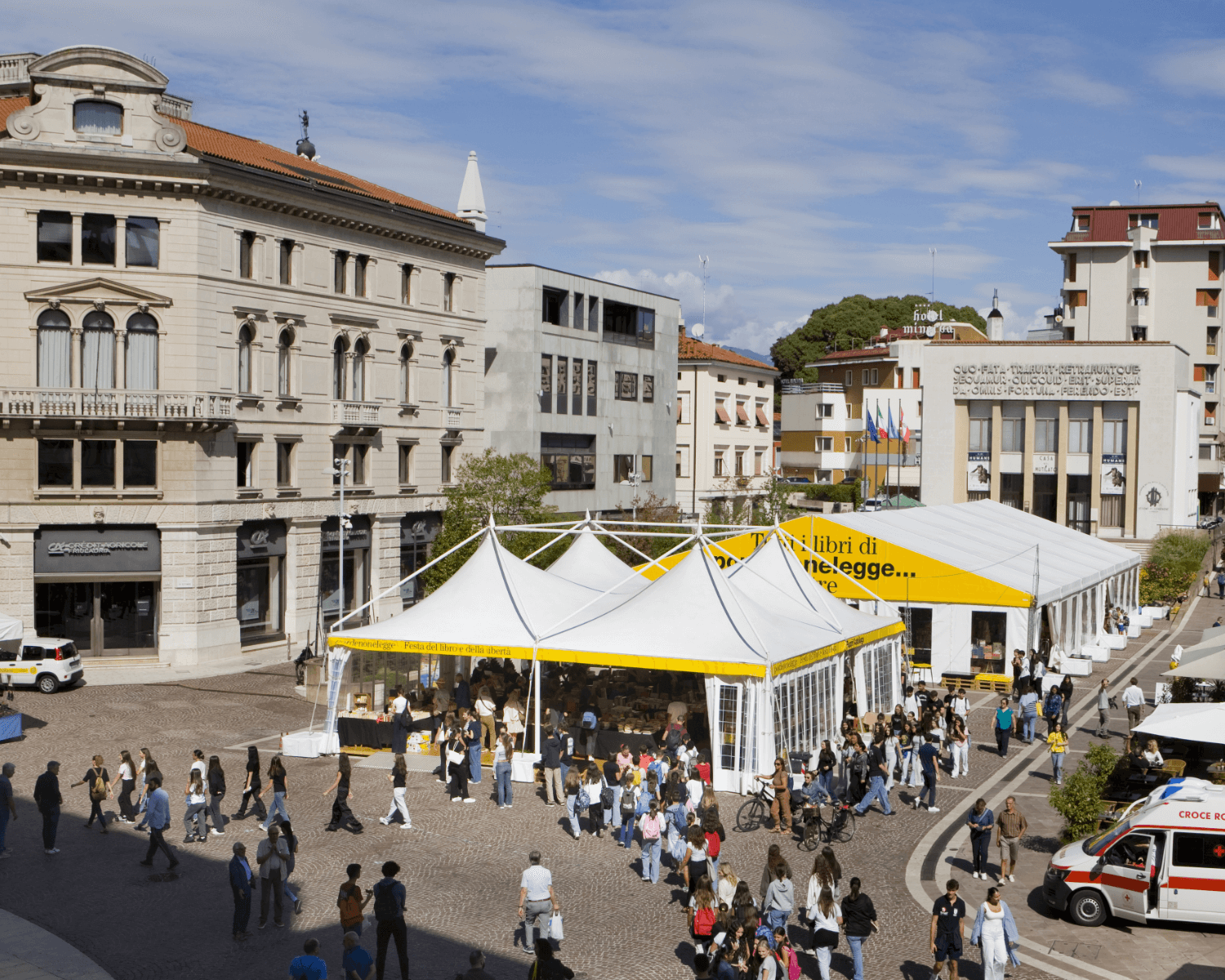 Una vivace piazza cittadina ospita una grande fiera del libro all'aperto sotto tendoni bianchi. Le persone camminano e si riuniscono intorno ai tendoni, mentre gli edifici storici e moderni circostanti fiancheggiano la piazza.