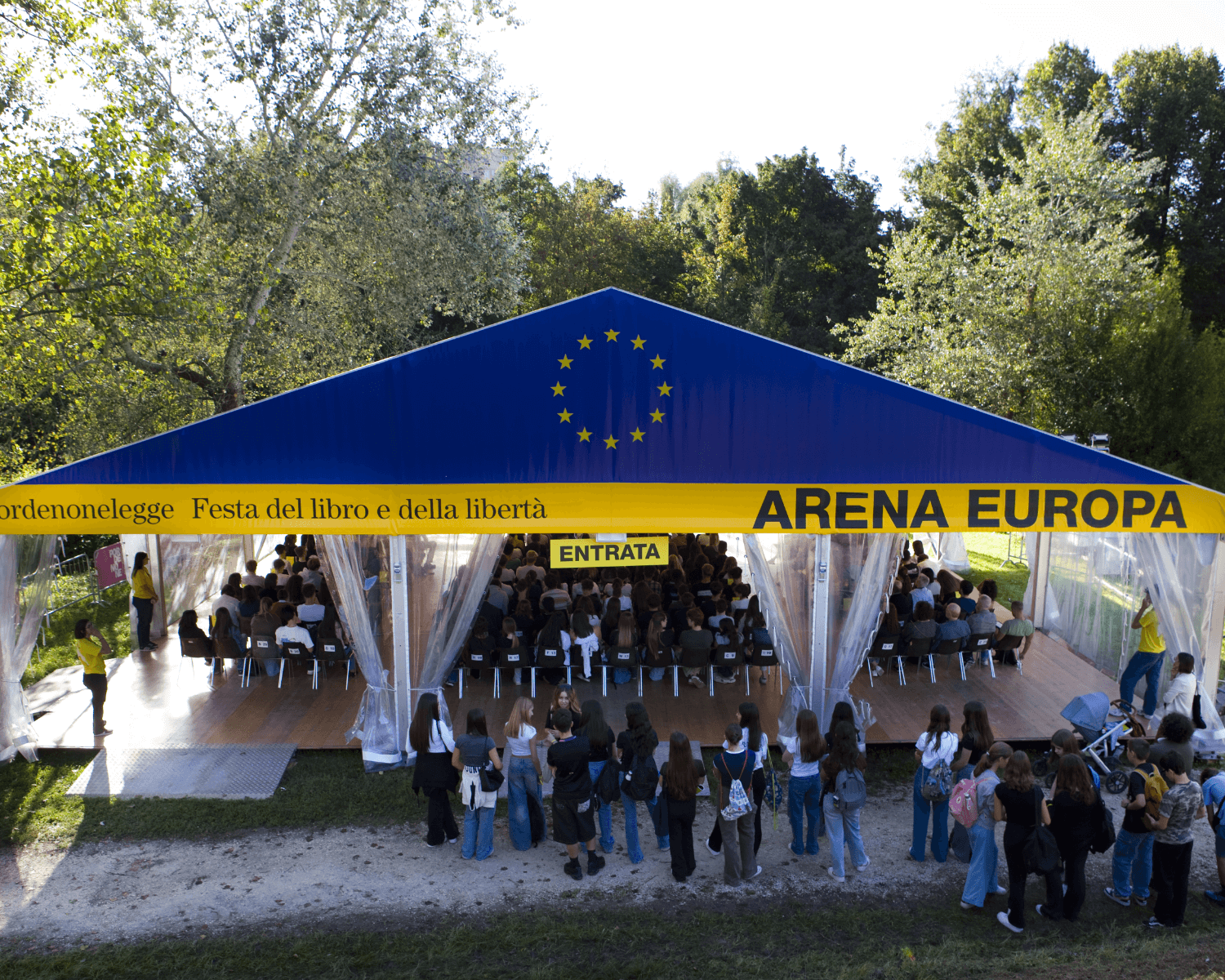 Un grande tendone blu e giallo con la scritta "ARENA EUROPA" ospita un pubblico seduto a un evento all'aperto, mentre le persone sono in fila all'esterno. Alberi e verde circondano l'area.
