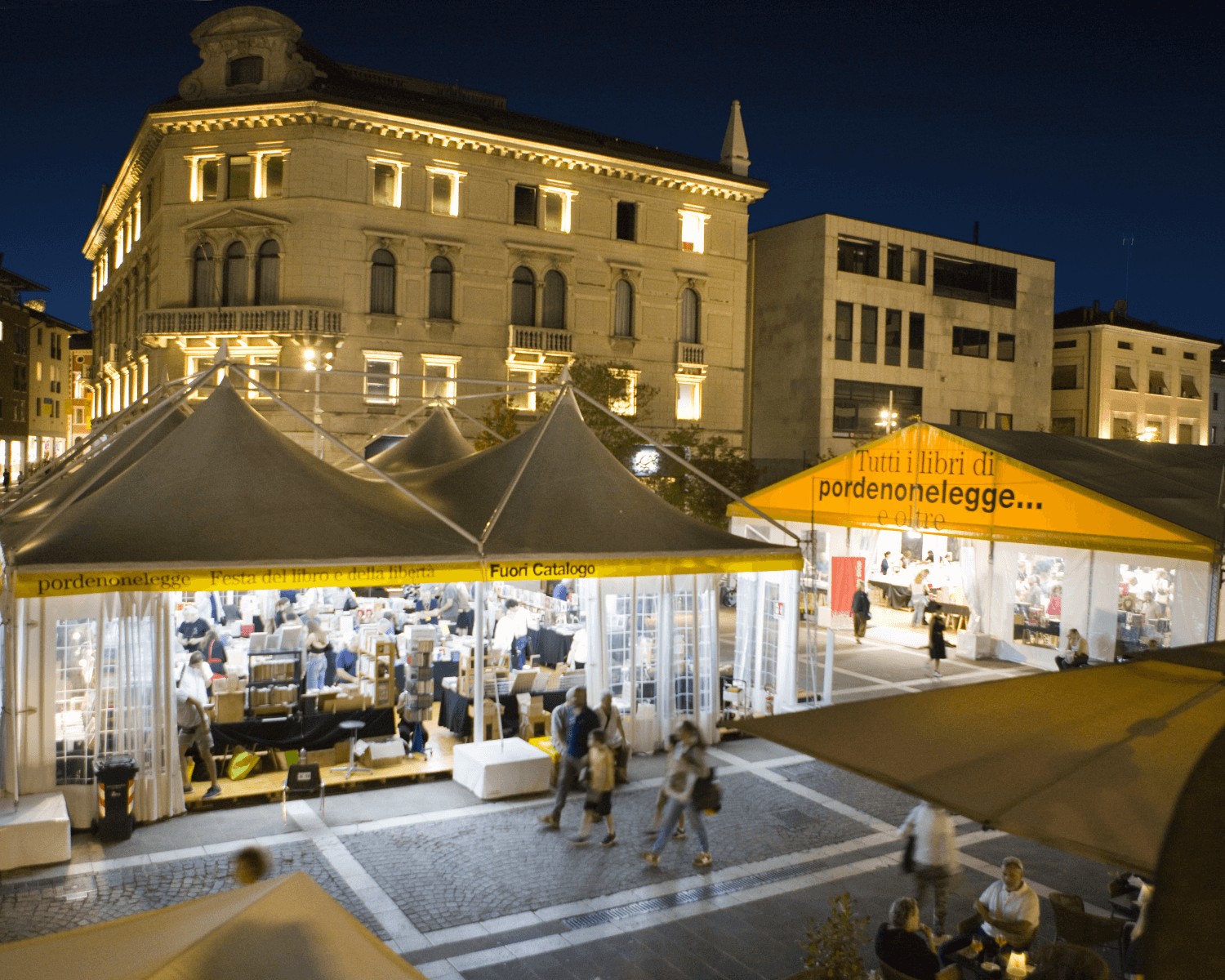 Una fiera del libro all'aperto illuminata di notte in una piazza di una città italiana, con persone che camminano e curiosano tra le bancarelle sotto grandi tendoni ed edifici storici illuminati sullo sfondo.