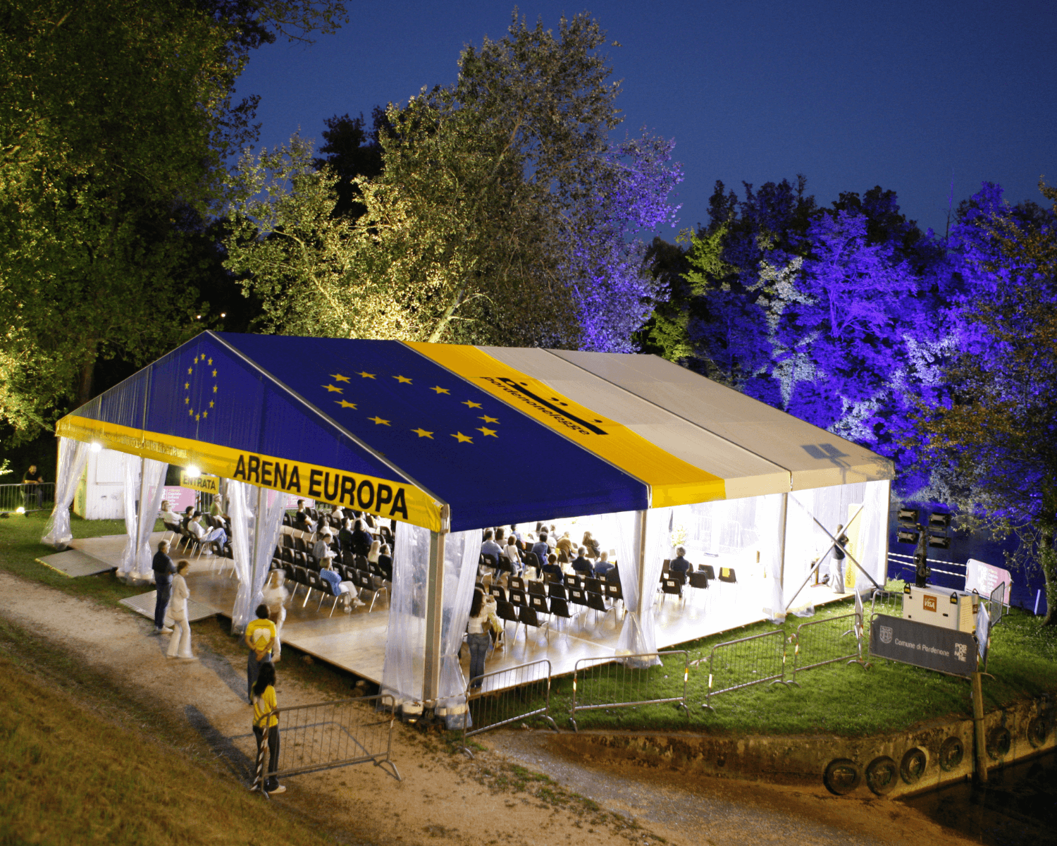 Una grande tenda all'aperto con la scritta "ARENA EUROPA", con i colori della bandiera dell'UE e le stelle sul tetto, allestita su un'area erbosa di notte. All'interno ci sono file di sedie e gli alberi intorno sono illuminati con luci colorate.
