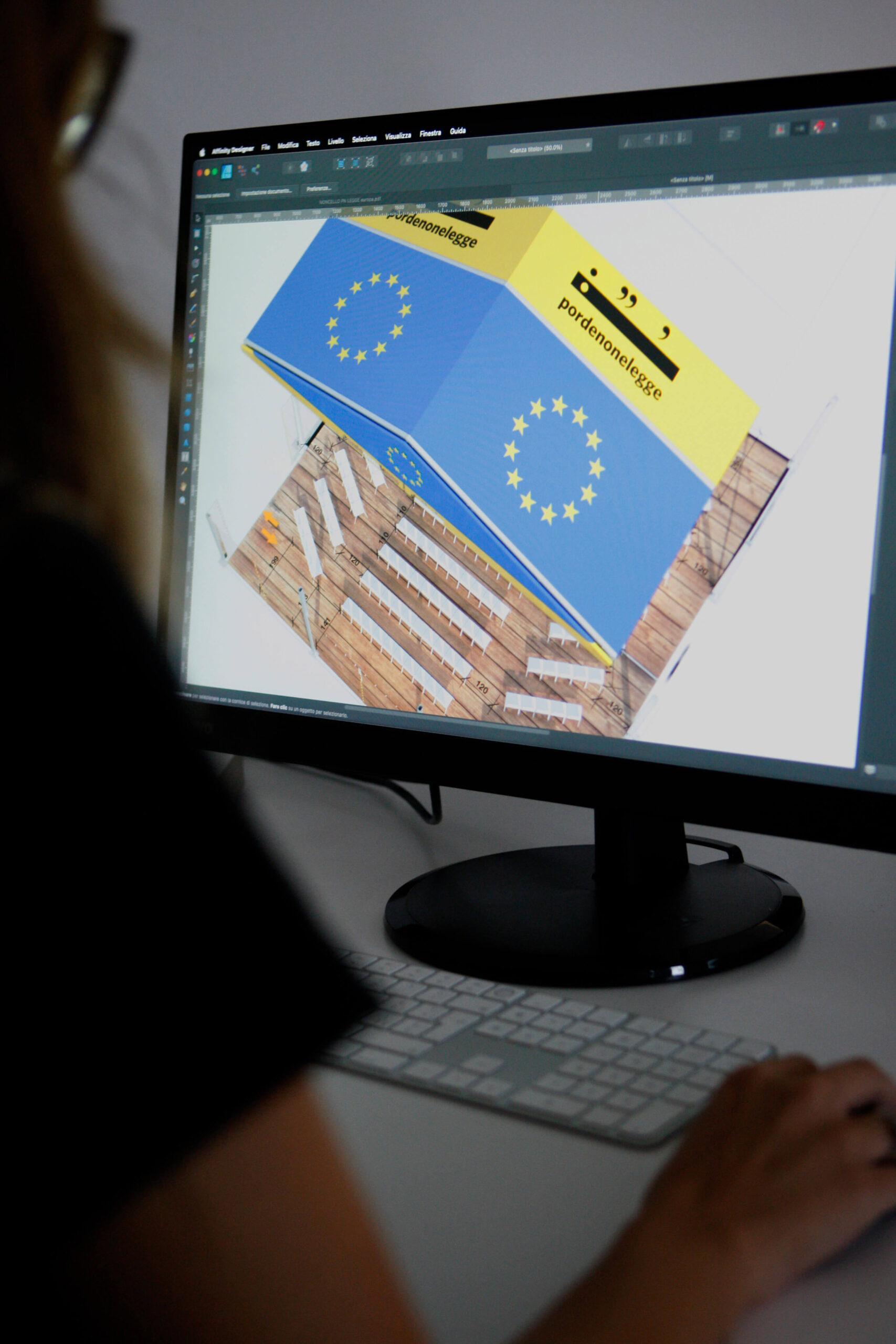 Una persona lavora al computer con un programma di progettazione aperto, visualizzando il modello 3D di una struttura con la bandiera dell'Unione Europea e il testo "proudtomeetalogue".