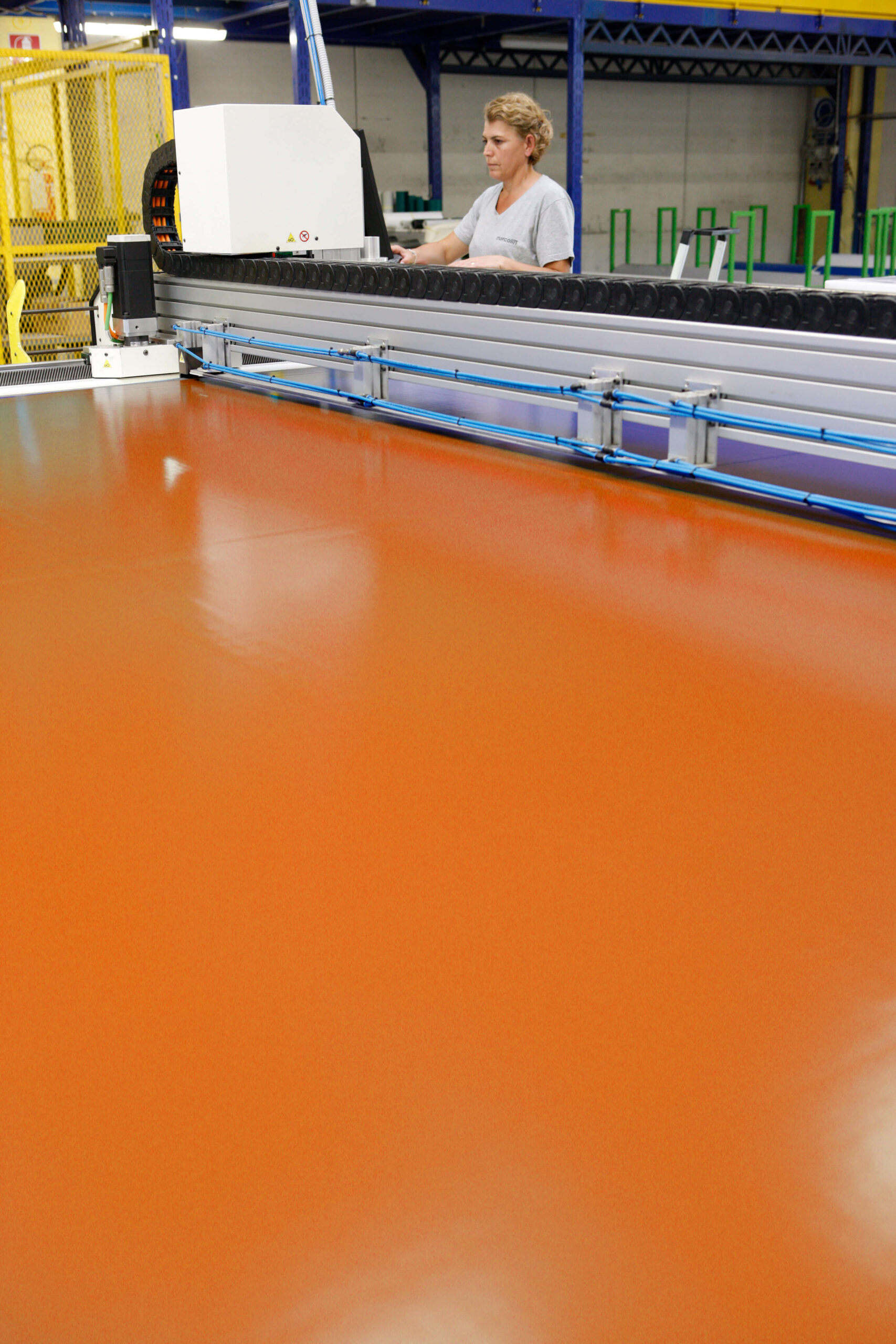 Una persona aziona una grande macchina industriale che lavora un ampio foglio di materiale arancione all'interno di una fabbrica con strutture metalliche e macchinari sullo sfondo.