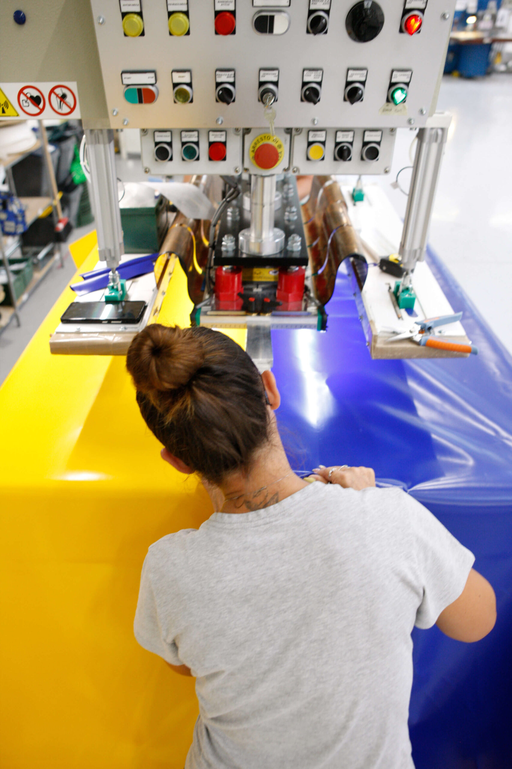 Una persona aziona una grande macchina industriale che unisce fogli di plastica gialli e blu in un ambiente industriale. La persona ha i capelli castani raccolti in uno chignon e indossa una camicia grigia.