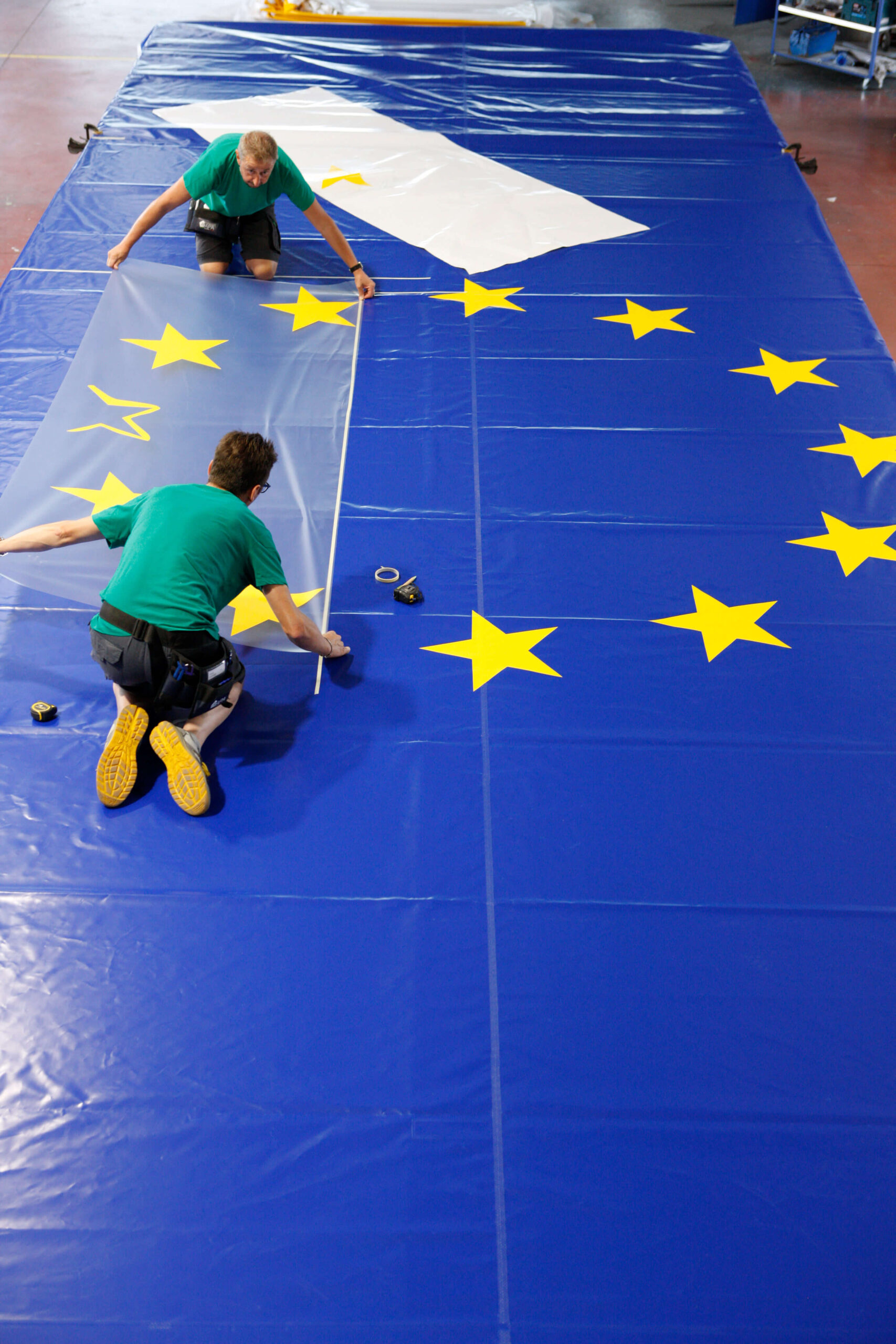 Due persone si inginocchiano su una grande bandiera dell'Unione Europea, sistemando un telo trasparente sulla sua superficie. La bandiera blu ha stelle gialle in cerchio e vari strumenti sono sparsi nelle vicinanze.