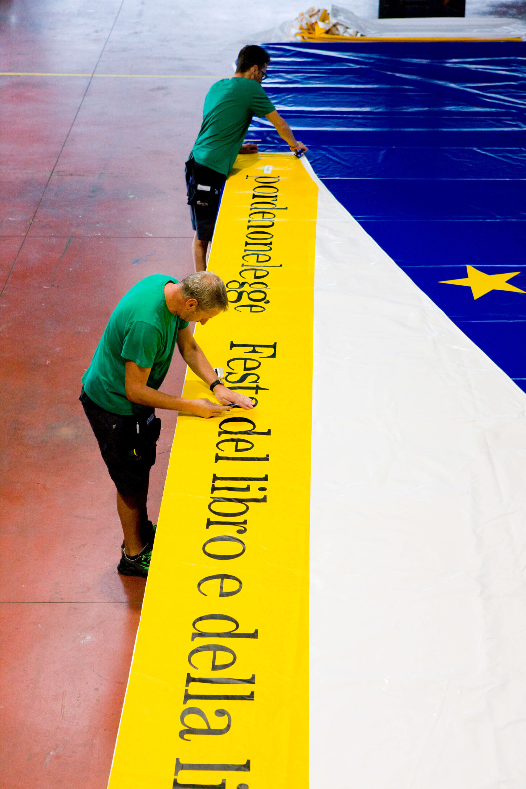 Due persone in camicia verde lavorano a un grande striscione con la scritta in italiano "Fest del Libro e della", accanto a una sezione blu con stelle gialle, al chiuso su un pavimento rossastro.