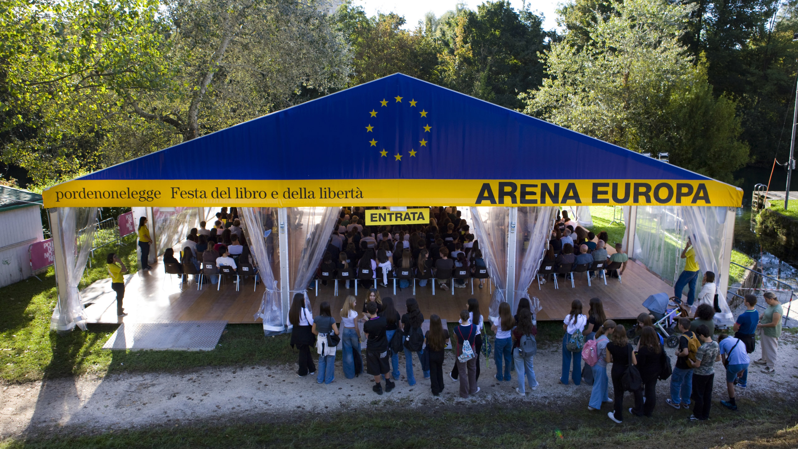 Grande tenda all'aperto con la scritta "ARENA EUROPA" e la bandiera dell'Unione Europea, piena di persone sedute. Una folla attende fuori per entrare. Lo striscione recita "pordenonelegge Festa del libro e della libertà". Alberi circondano l'area.