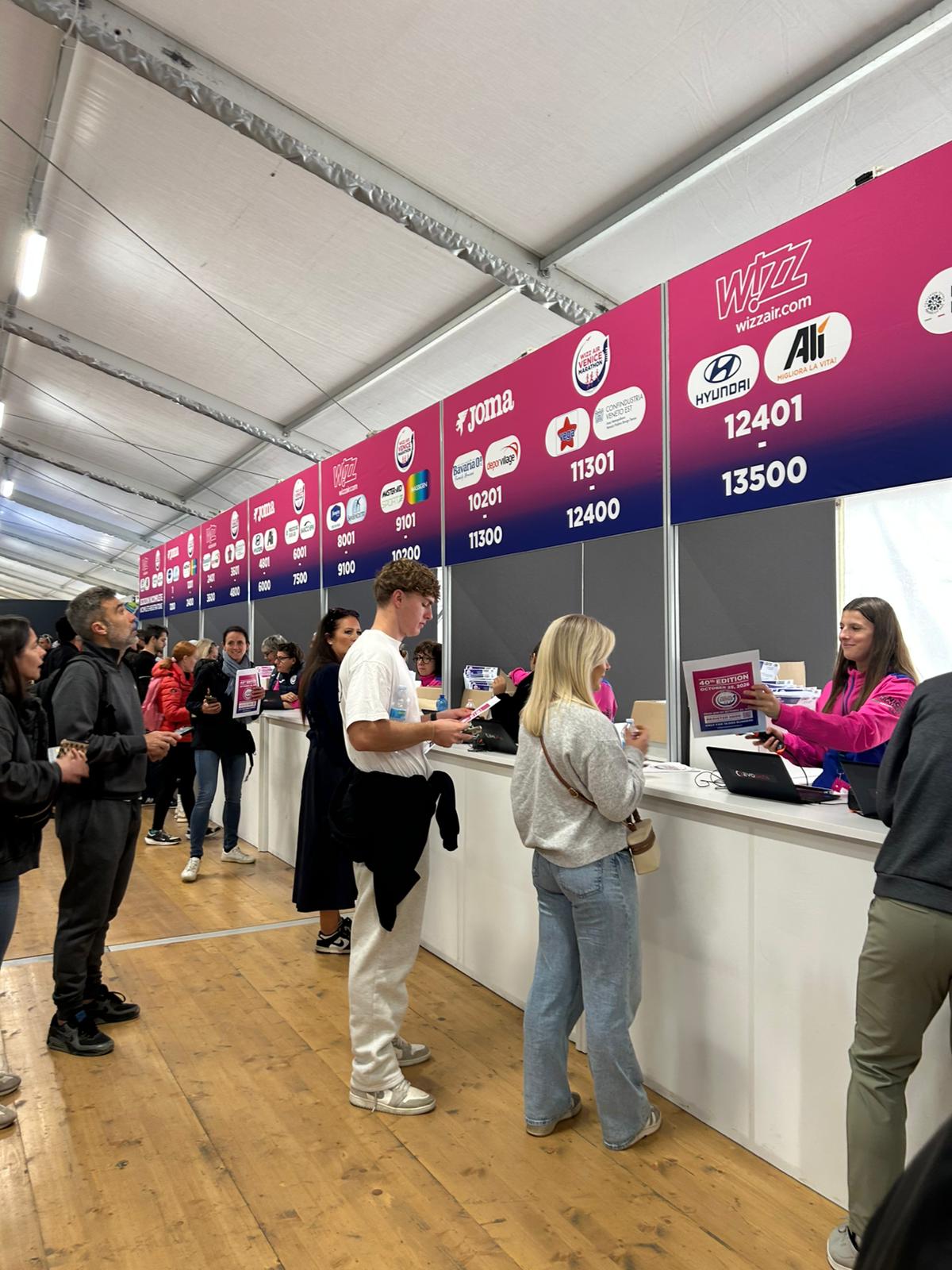 Le persone sono in fila al banco di registrazione all'interno di una grande tenda e ricevono il materiale dal personale dietro le scrivanie. I cartelli sopra ogni banco mostrano i loghi degli sponsor e le fasce numeriche per il check-in dei partecipanti.