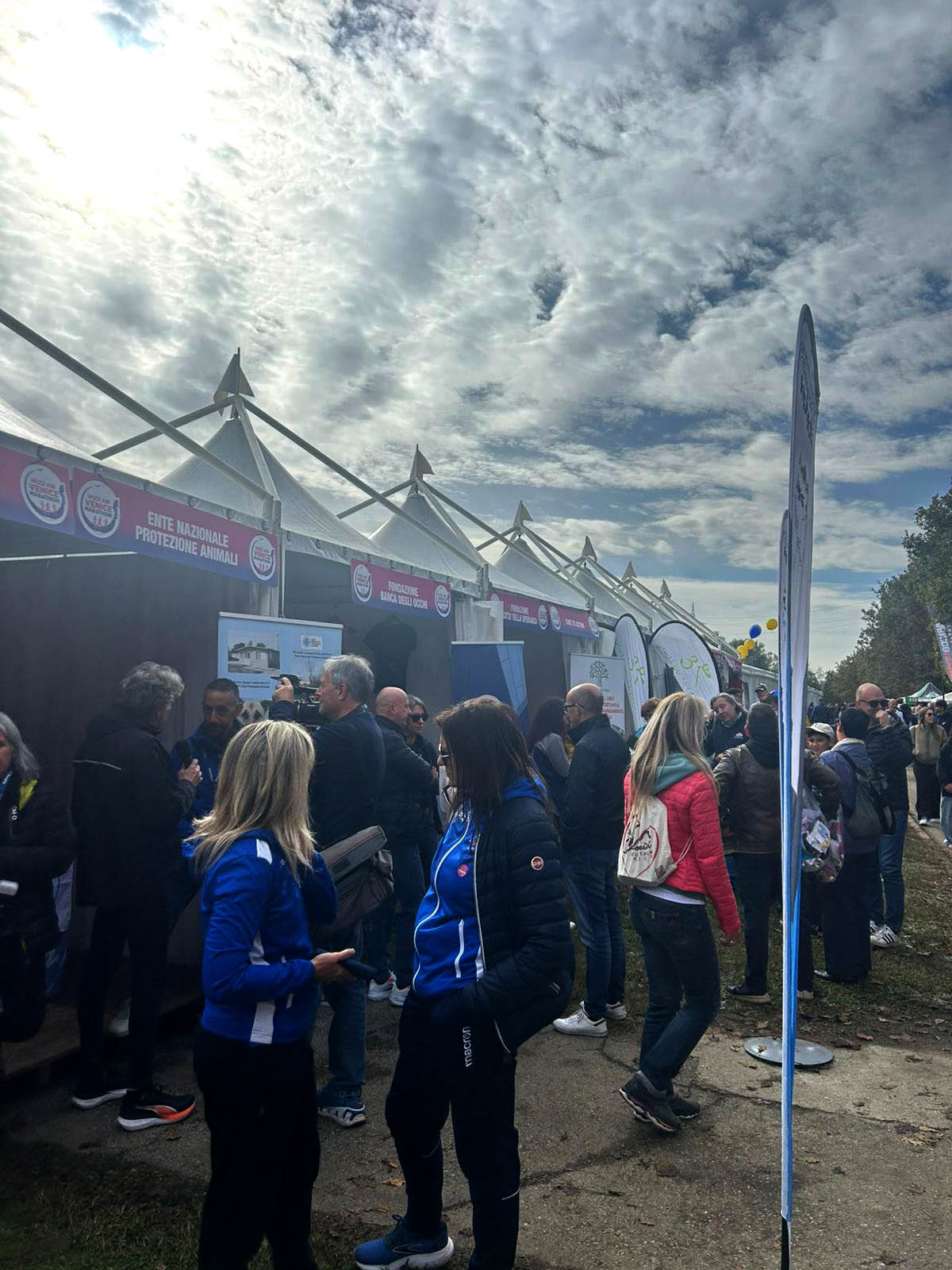Le persone si radunano e si mettono in fila davanti a una fila di tende bianche in occasione di un evento all'aperto, con un cielo nuvoloso in testa. Alcuni indossano giacche blu e sopra i tendoni sono visibili cartelli e striscioni informativi.