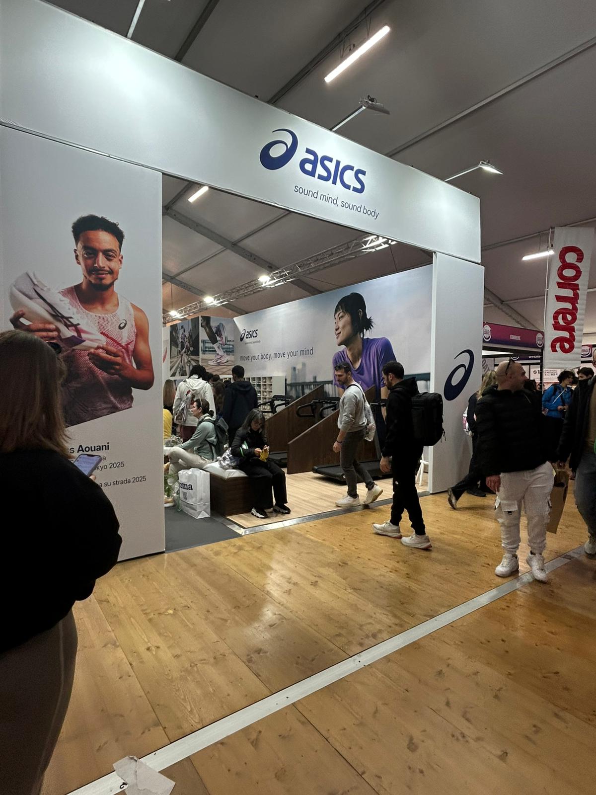 Un affollato stand Asics a un evento indoor presenta grandi poster di atleti, scaffali con scarpe e persone che camminano e interagiscono con gli oggetti esposti su un pavimento di legno.