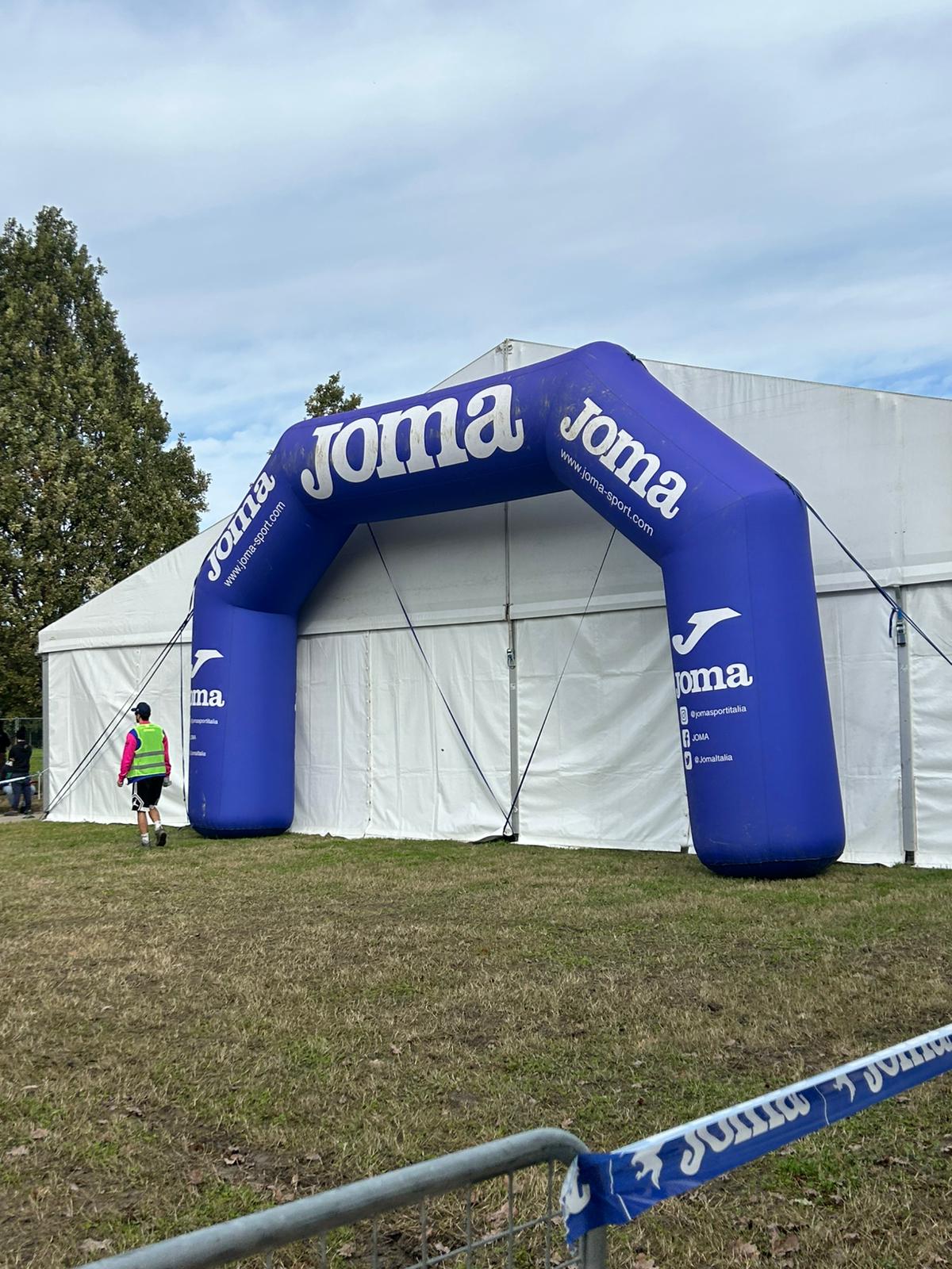 Una persona in tenuta sportiva si dirige verso un grande arco gonfiabile blu con il marchio "Joma", allestito accanto a una tenda bianca su un'area erbosa, con alberi e un cielo nuvoloso sullo sfondo.