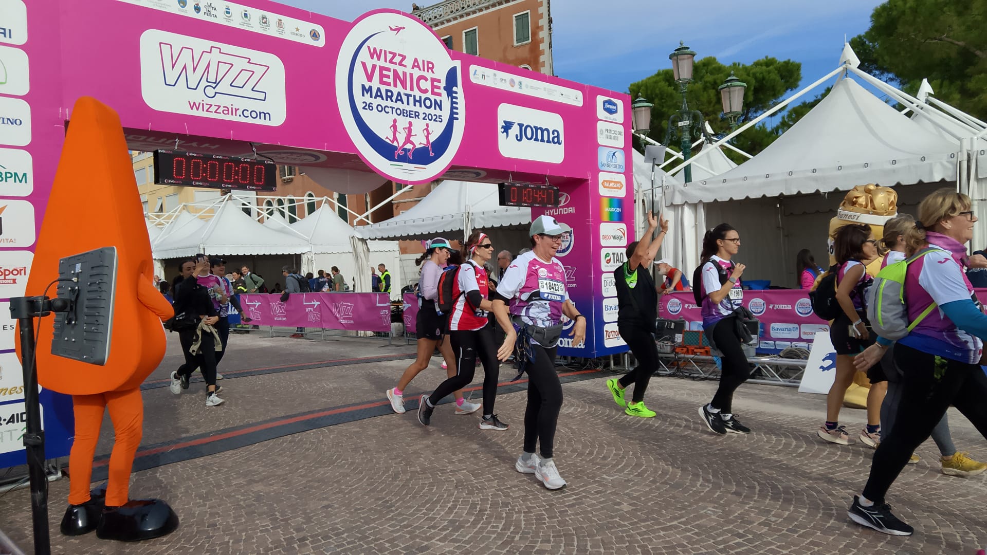 Runners cross the finish line at the Wizz Air Venice Marathon 2025, passing under a pink arch surrounded by event banners. People are wearing race bibs and sportswear, with some waving and smiling.