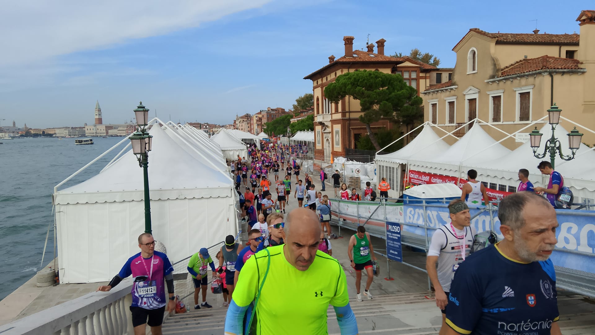 Corridori partecipano a una corsa lungo un canale di Venezia, con tende bianche che costeggiano il lungomare ed edifici storici sullo sfondo. Alcuni corridori salgono su gradini di pietra in primo piano. Il cielo è parzialmente nuvoloso.