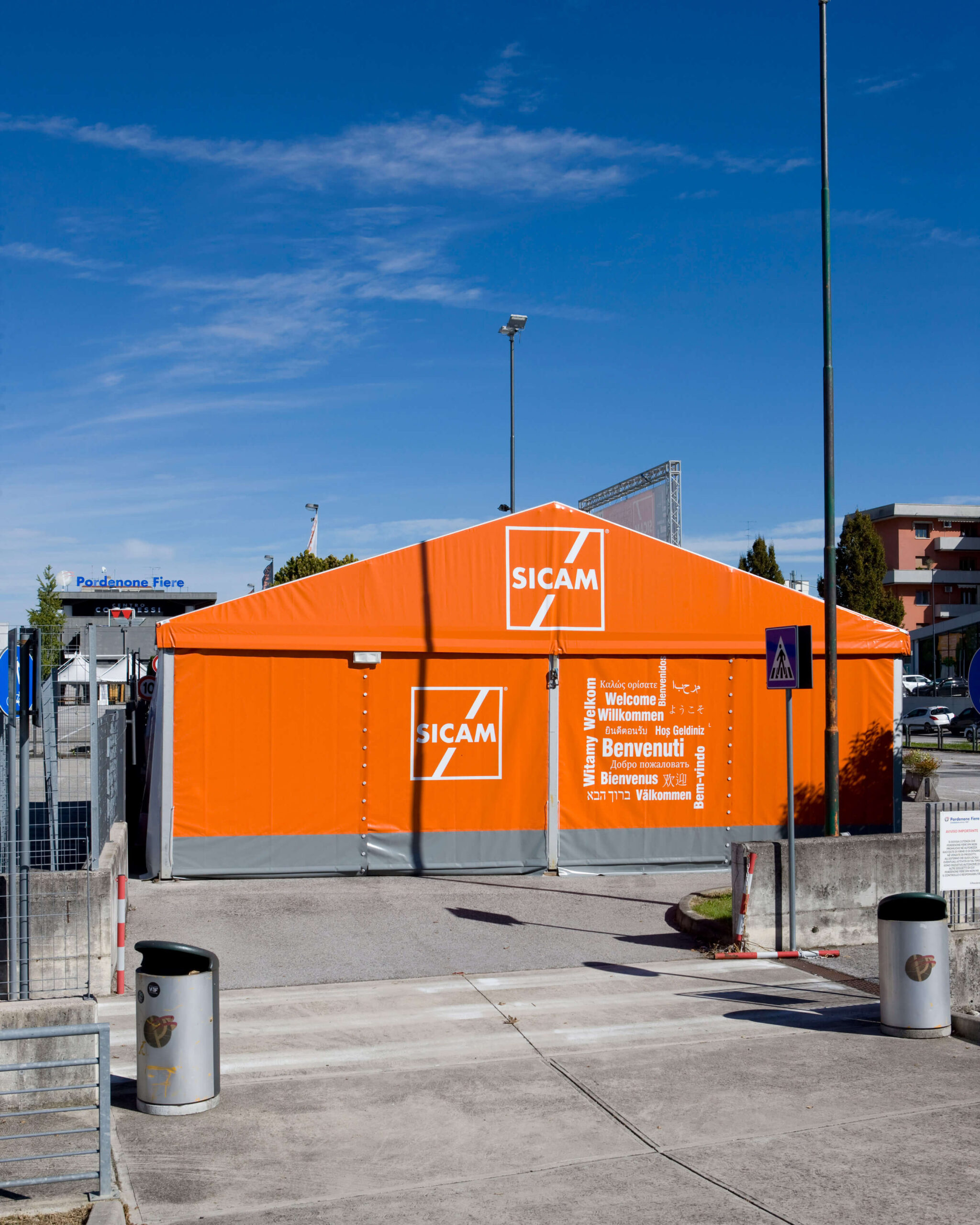 Una grande tenda arancione con la scritta "SICAM" si trova all'ingresso dell'evento. Sulla facciata sono visibili cartelli di benvenuto in diverse lingue. Cestini e transenne delimitano il percorso asfaltato che conduce alla tenda.