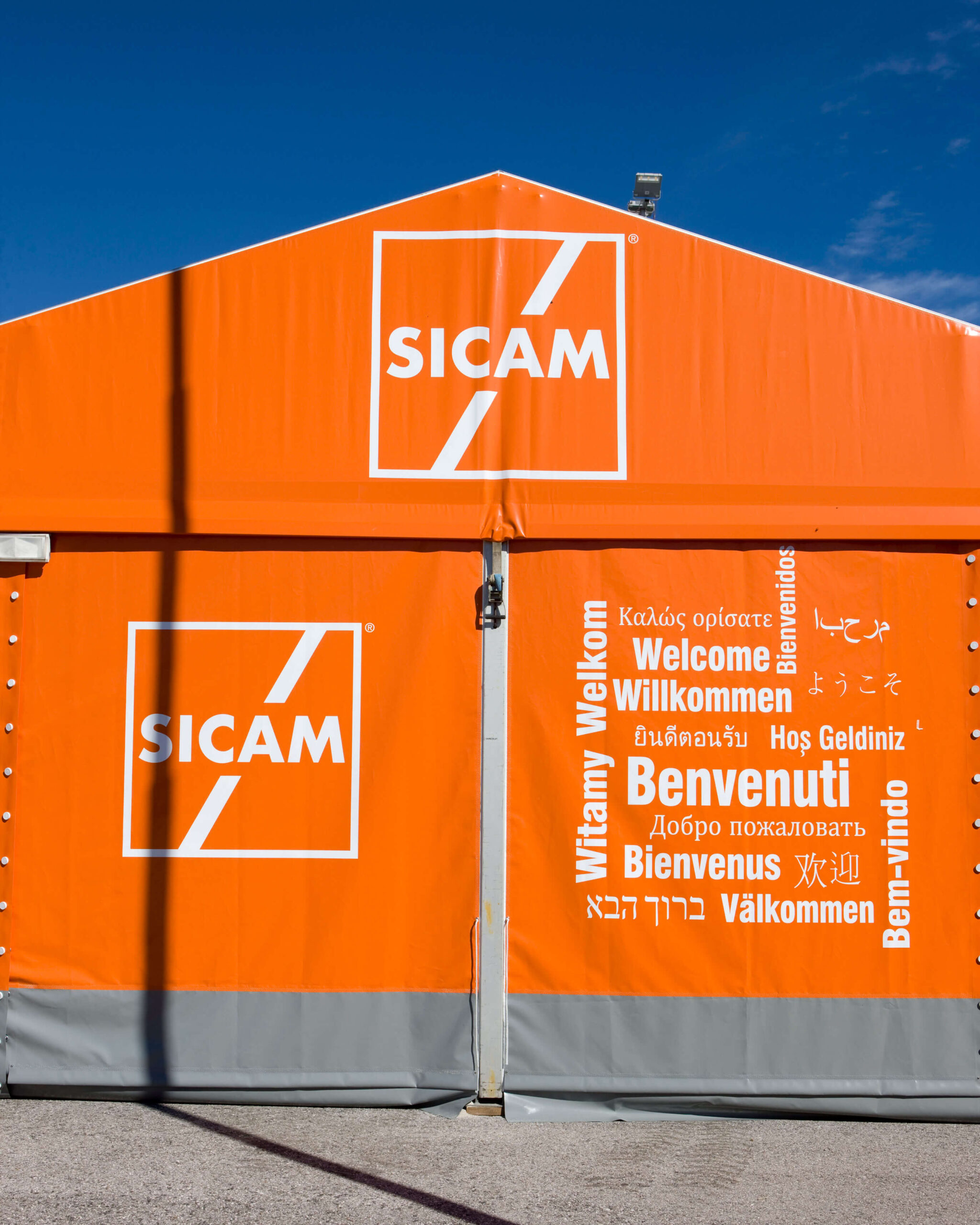 Una tenda arancione con il logo SICAM e la parola "Welcome" scritta in più lingue, sullo sfondo di un cielo azzurro e limpido.
