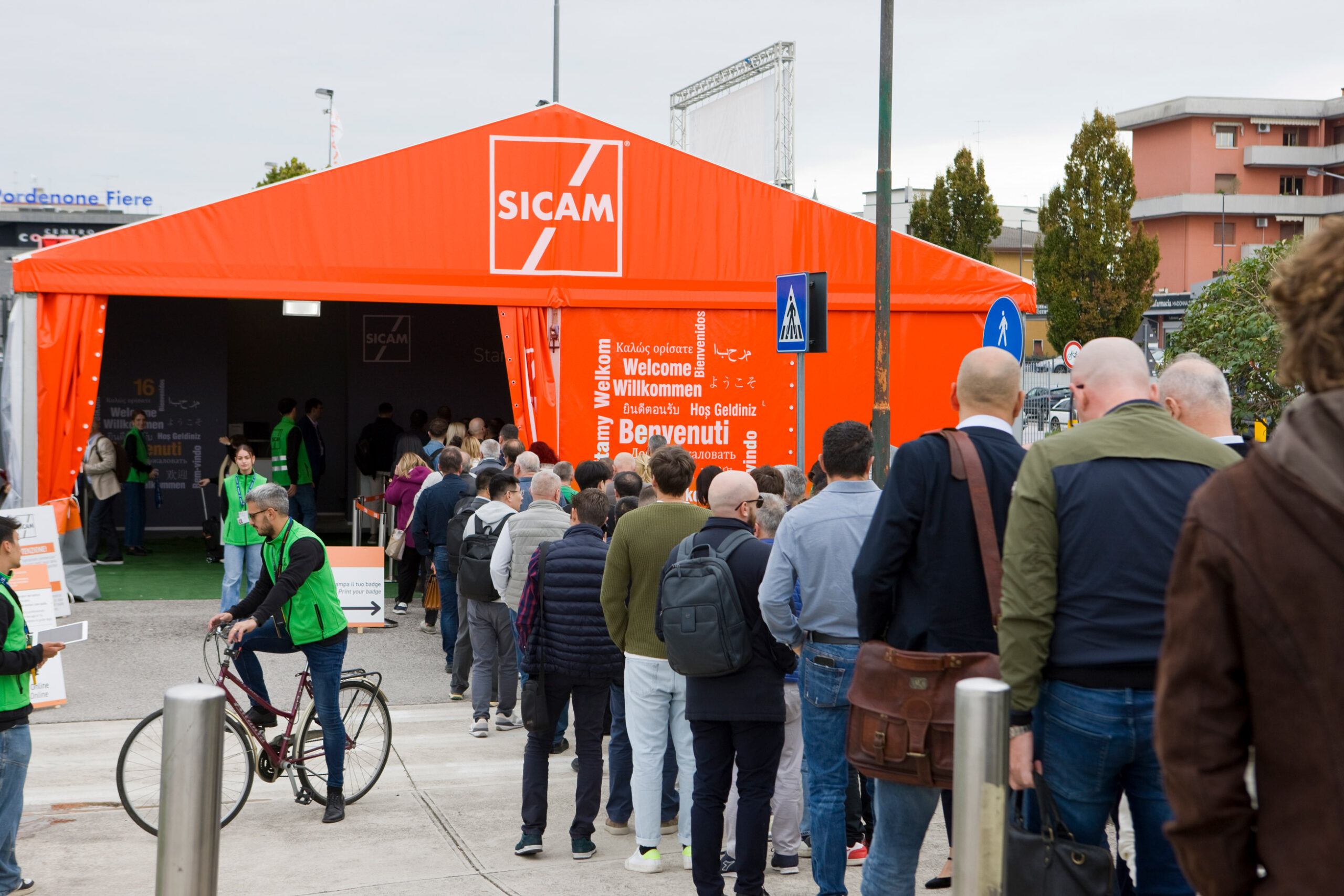 Una lunga fila di persone attende di entrare nel grande tendone arancione della SICAM. Alcuni membri dello staff in gilet verde assistono i partecipanti vicino all'ingresso. Un ciclista passa sul marciapiede. Sullo sfondo sono visibili edifici e alberi.