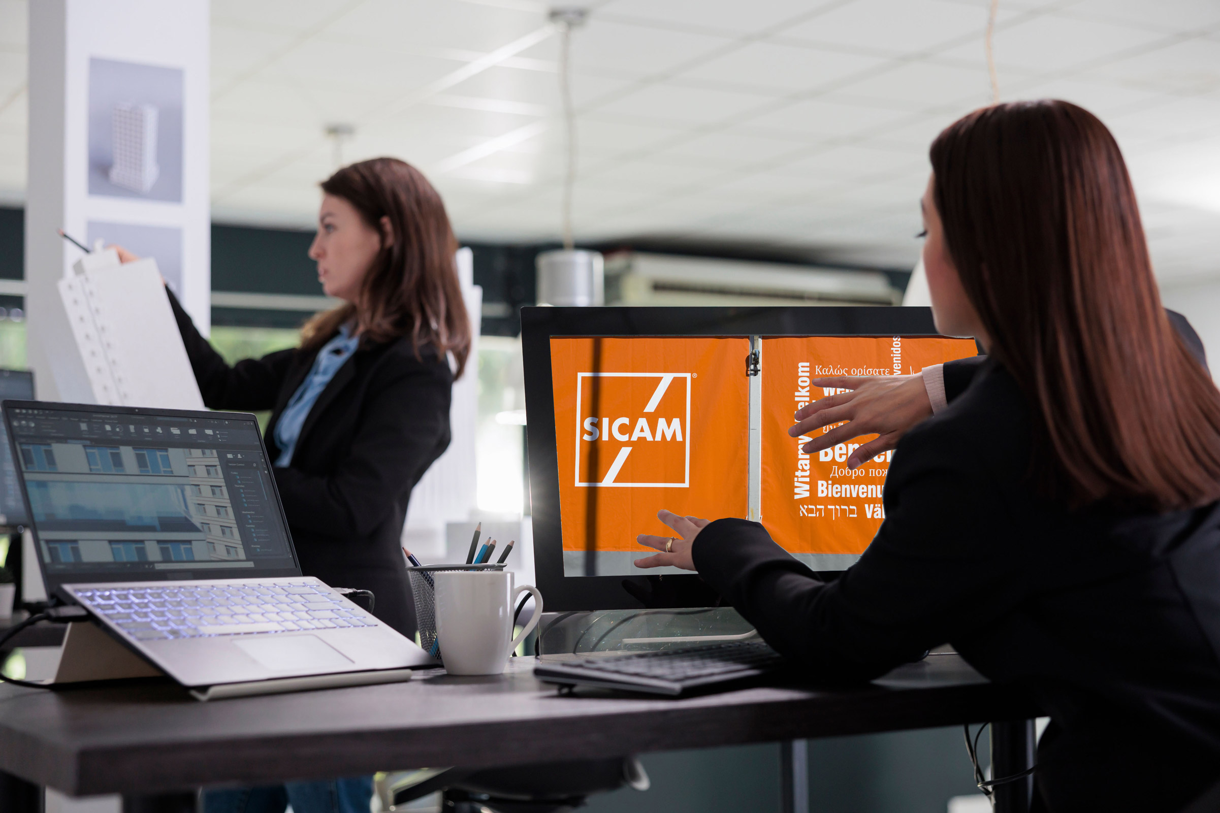 Due donne lavorano in un ufficio moderno. Una esamina dei documenti, mentre l'altra gesticola verso lo schermo di un computer che mostra una presentazione arancione con la scritta "SICAM". Sulla scrivania sono presenti un computer portatile, una tazza da caffè e altri articoli da ufficio.