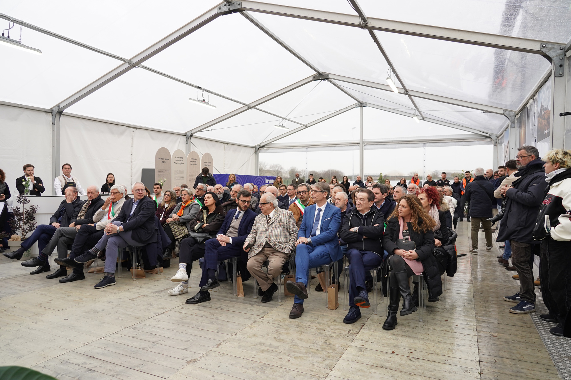 Un folto gruppo di persone, sedute e in piedi, assiste a un evento all'interno di una tenda con tetto trasparente. Il pubblico appare attento, vestito per lo più con abiti business o smart casual.