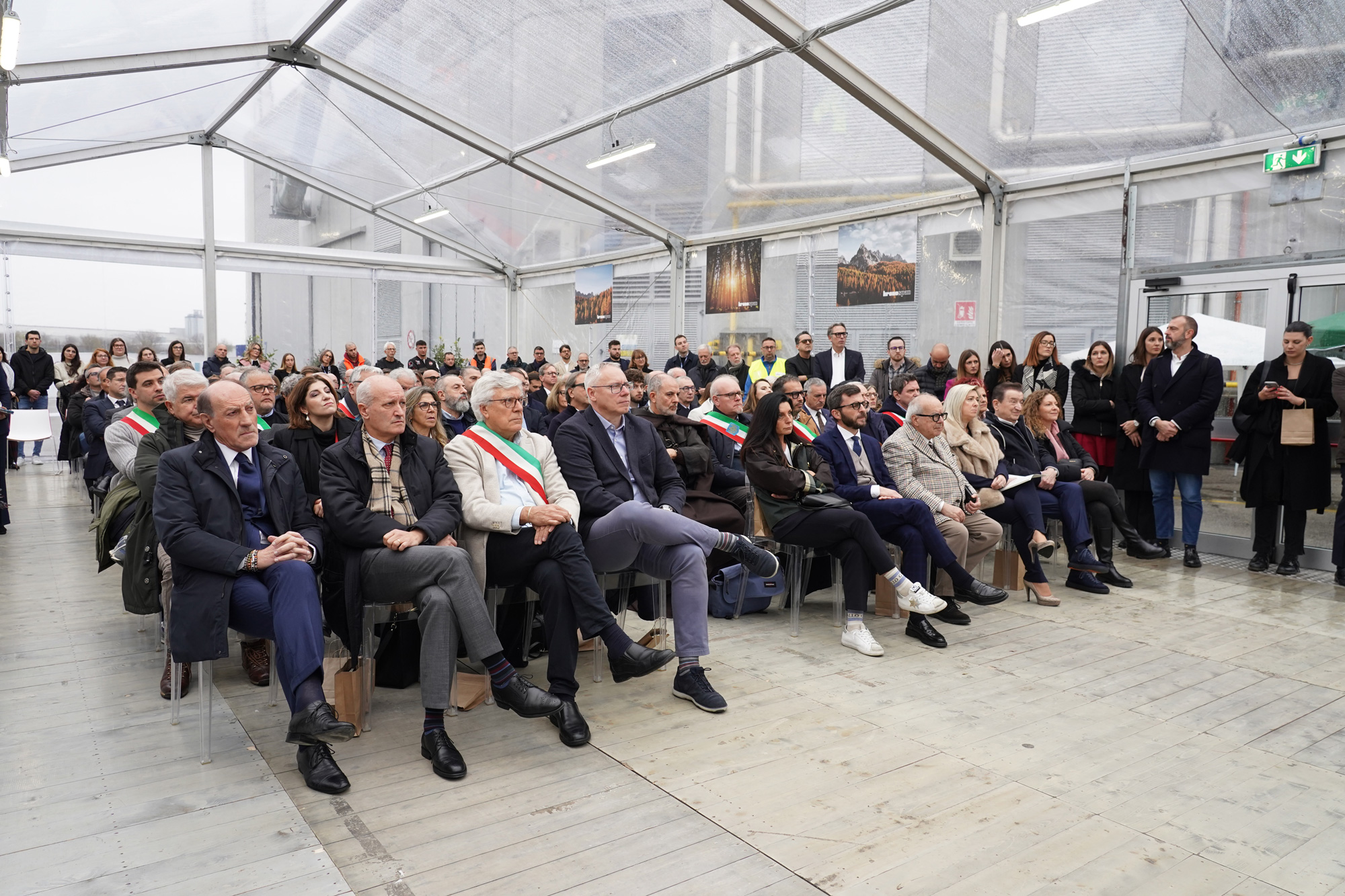 Un folto gruppo di persone, tra cui uomini e donne di varie età, siedono e stanno in piedi con attenzione sotto una tenda trasparente durante un evento formale. Alcune persone sedute indossano una fascia che suggerisce un ruolo ufficiale.