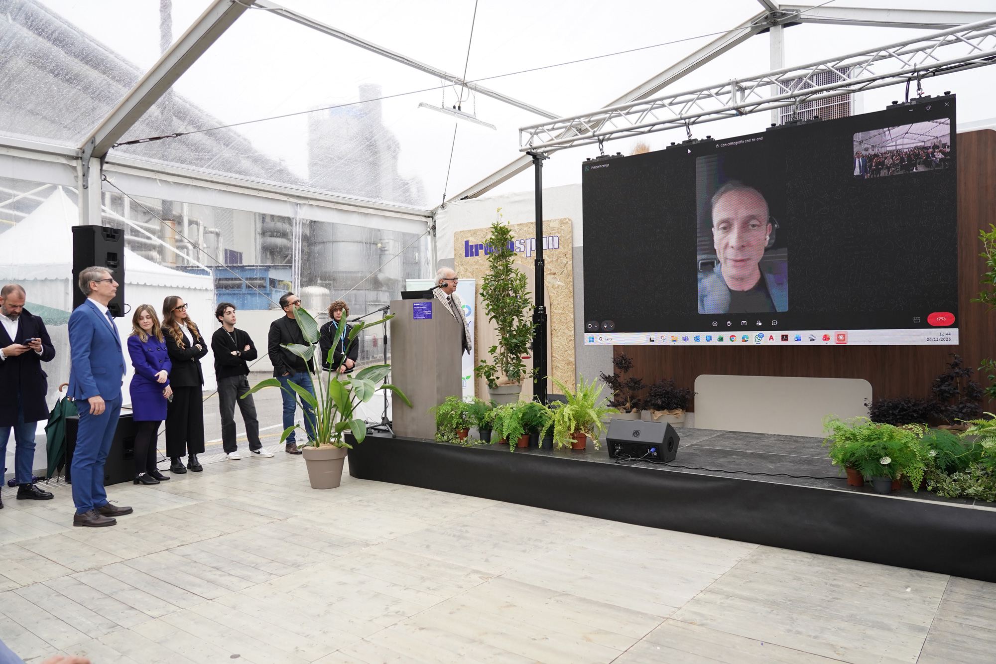 Un gruppo di persone si trova sotto una grande tenda e ascolta un oratore su un podio. Un grande schermo mostra un uomo che partecipa tramite videochiamata. L'area è decorata con piante verdi.