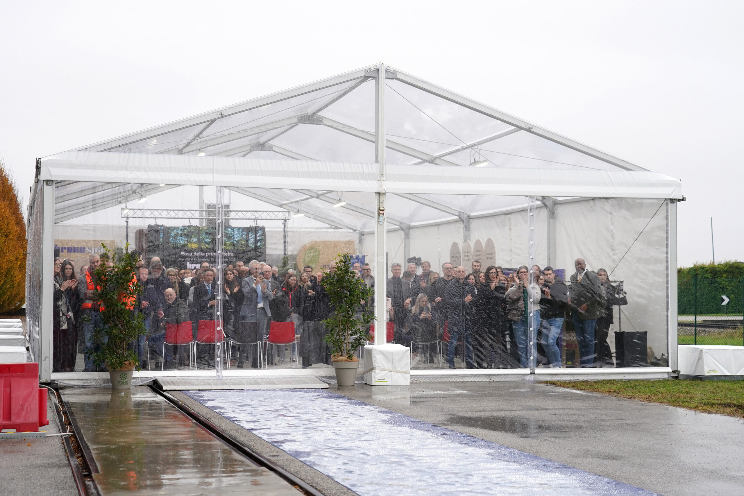 Un gruppo di persone si trova e si siede all'interno di una grande tenda trasparente durante una giornata di pioggia, partecipando a un evento all'aperto; le piante decorano l'ingresso e un sentiero bagnato conduce alla tenda.