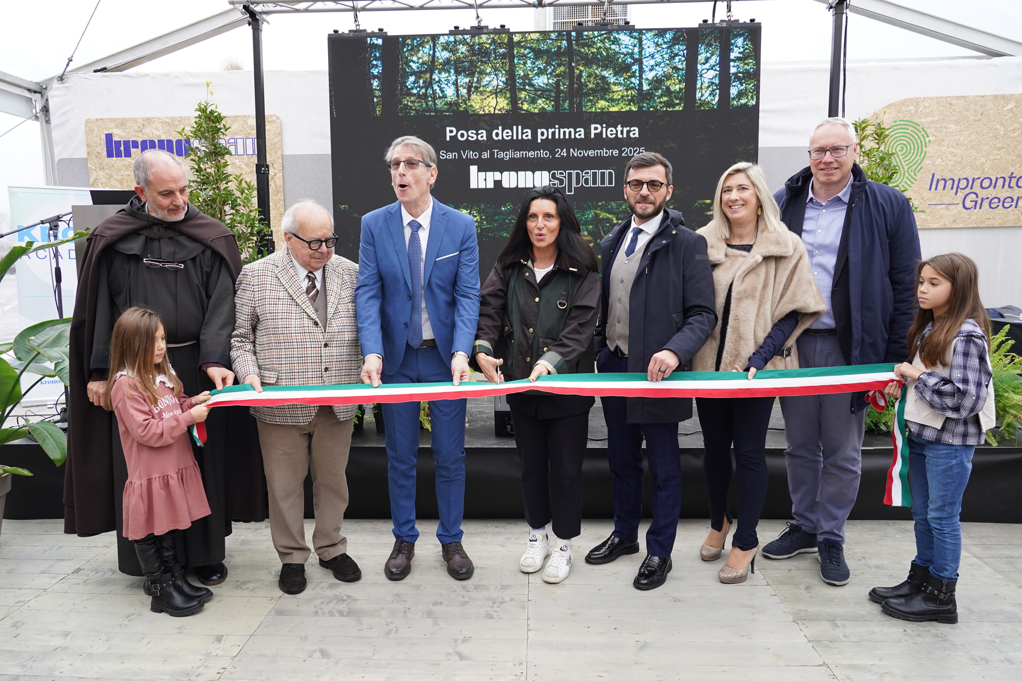 Otto persone, tra cui due bambini, sono in fila e tengono in mano un lungo nastro tricolore durante un evento. Dietro di loro c'è uno schermo con un testo in italiano e del verde, che indica una cerimonia o una celebrazione.