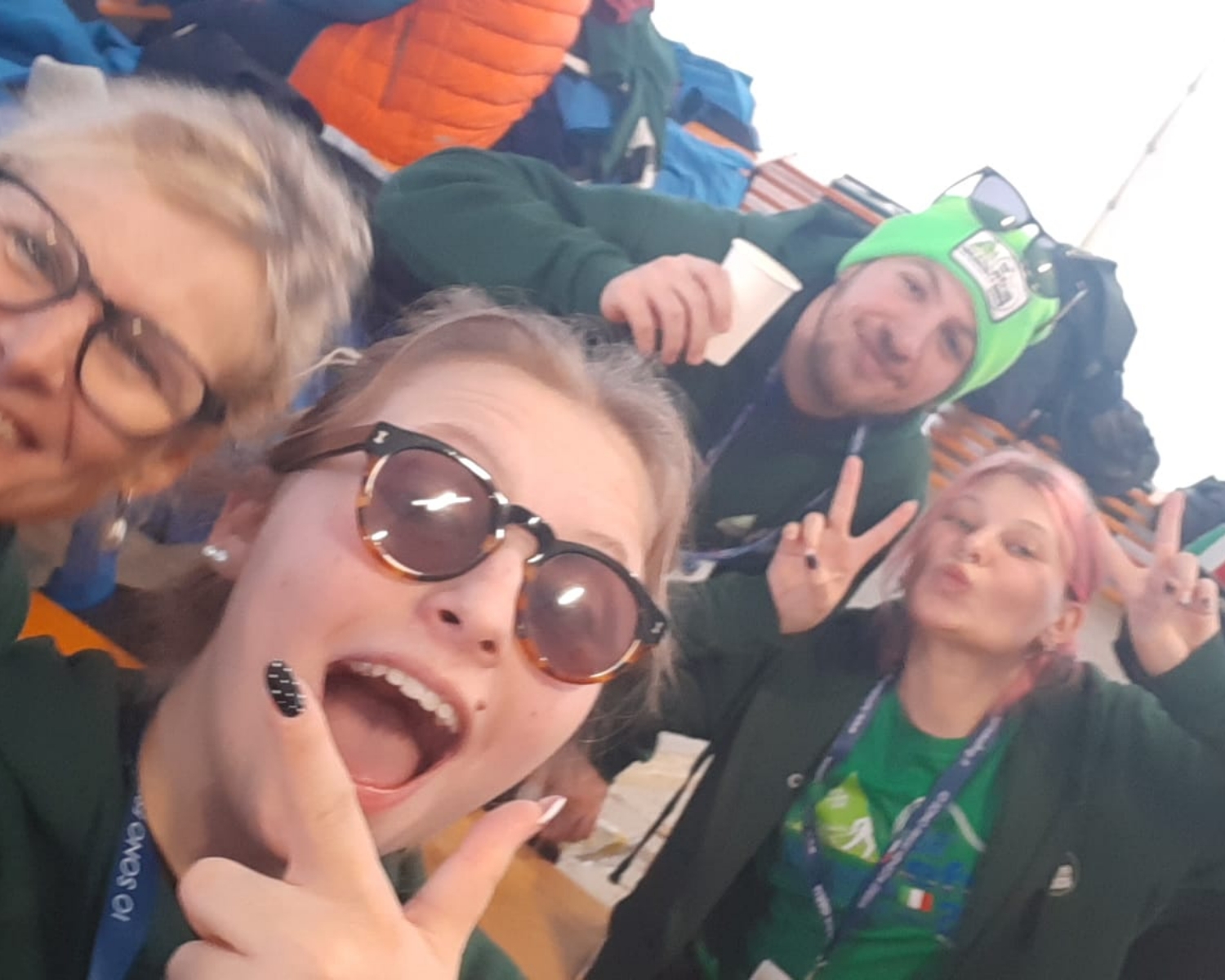 Quattro persone in abiti casual sorridono e posano per un selfie. Una persona indossa occhiali da sole e fa il segno della pace, un'altra indossa un berretto verde e altre due sorridono e gesticolano. Sullo sfondo ci sono borse colorate.