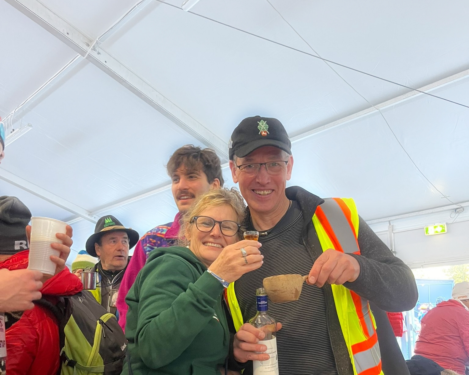 Una donna sorridente con gli occhiali abbraccia un uomo con giubbotto e berretto ad alta visibilità; entrambi tengono in mano dei drink. Attorno a loro, all'interno di una grande tenda, persone con giacche e cappelli colorati. L'atmosfera è festosa e allegra.