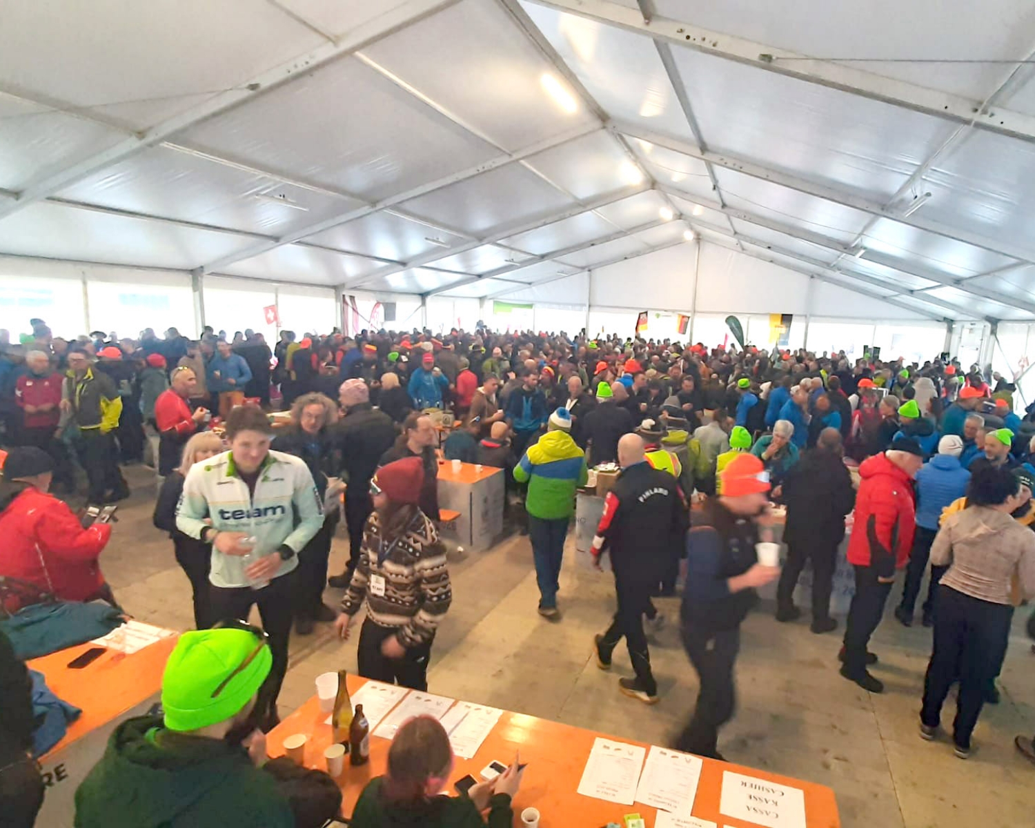 Una grande folla di persone con giacche e cappelli colorati si riunisce all'interno di una tenda spaziosa e ben illuminata con tavoli, alcuni in piedi e altri seduti, suggerendo un evento vivace o un'atmosfera da festival.