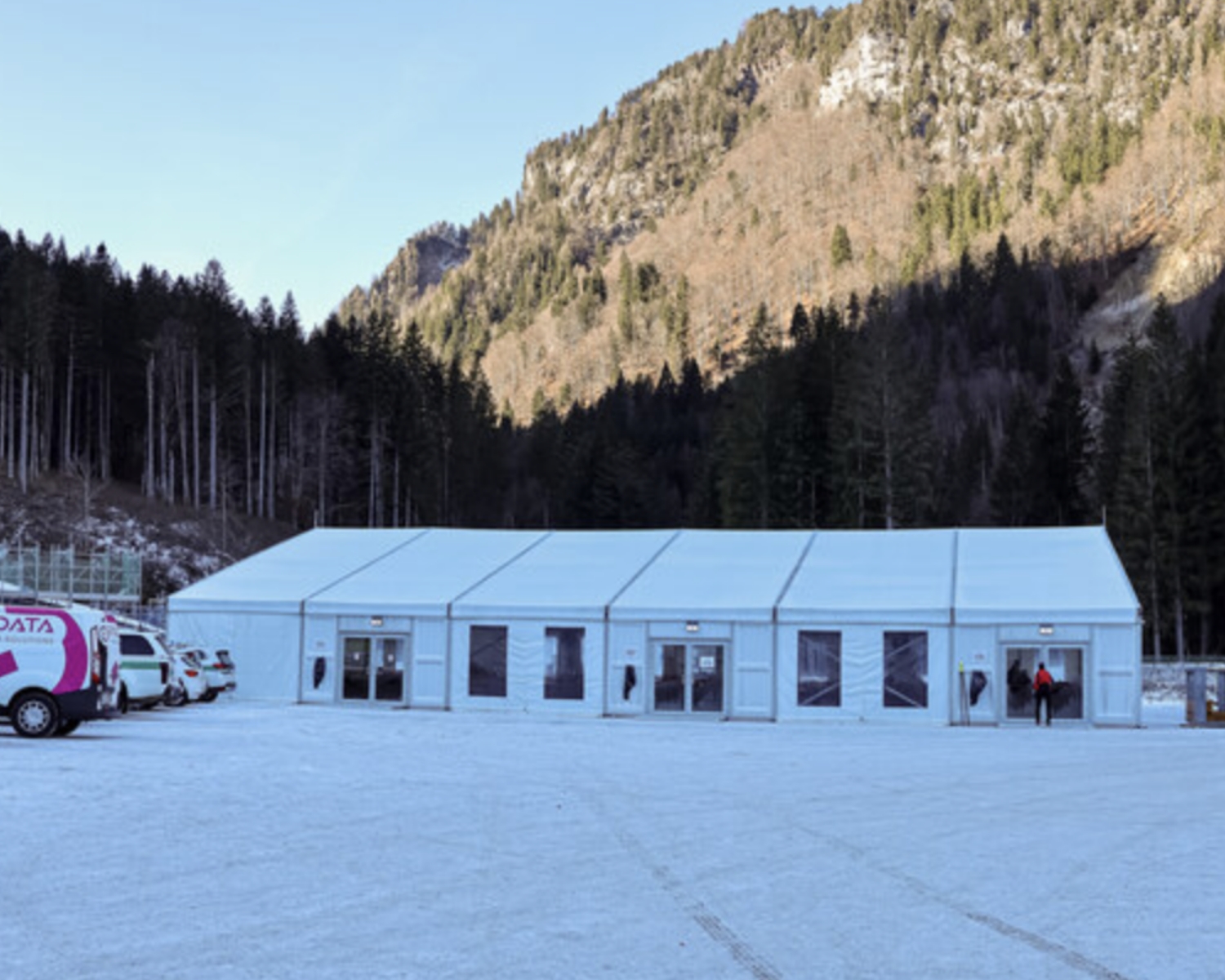 Una grande tenda bianca si trova su un terreno coperto di neve, con alcune persone e veicoli nelle vicinanze. Sullo sfondo si ergono montagne boscose sotto un cielo limpido.