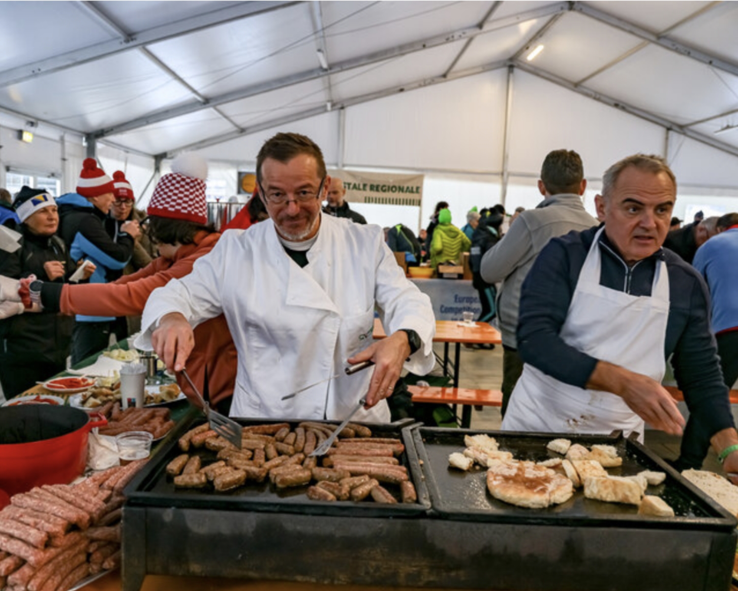 Due uomini in grembiule cucinano salsicce e carni su una grande griglia in un mercato al coperto. Sullo sfondo, persone in abiti invernali chiacchierano e aspettano il cibo sotto una tenda bianca.