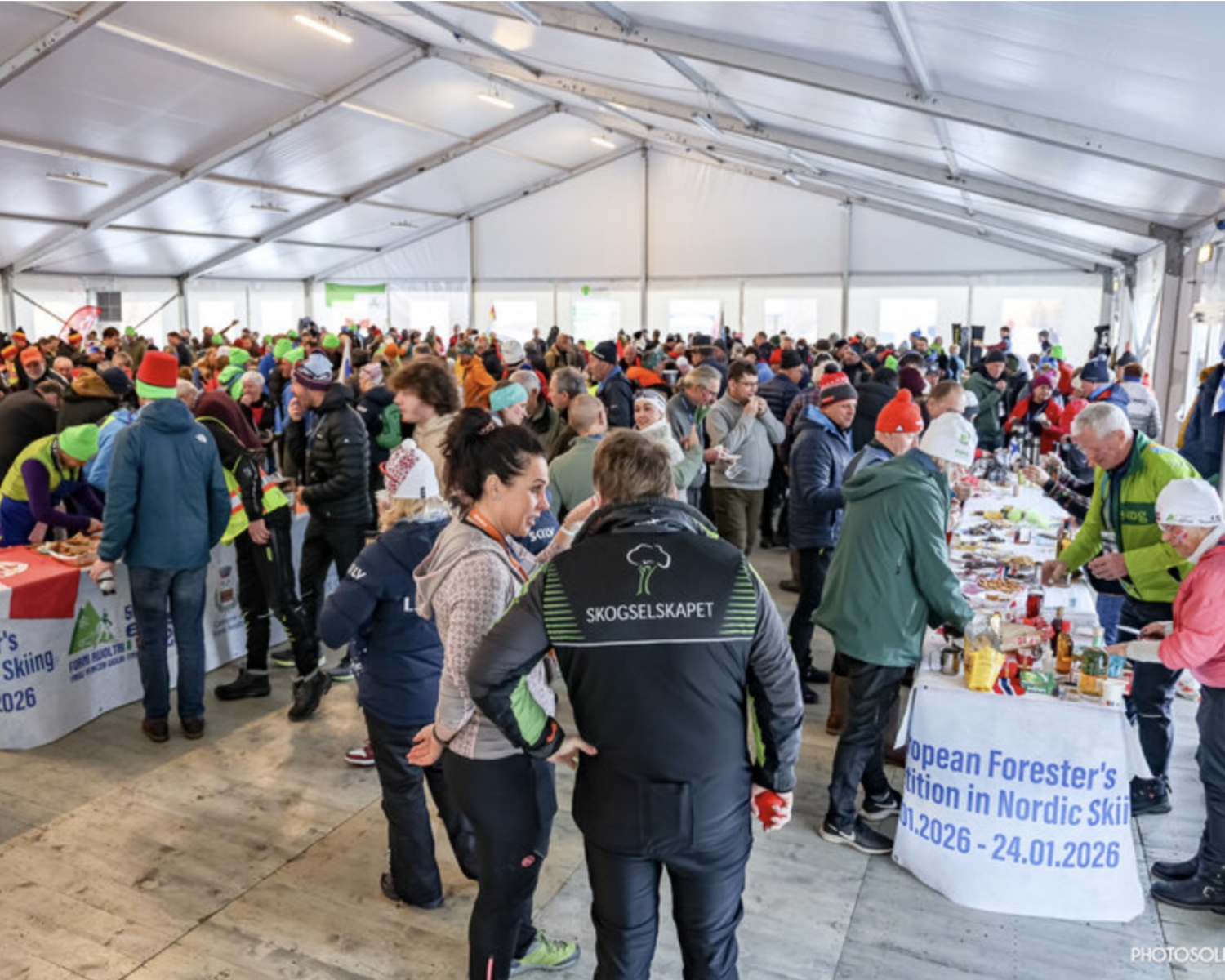 Un folto gruppo di persone si riunisce all'interno di un'ampia tenda per eventi con stand informativi, tavoli di ristoro e persone che indossano abbigliamento e cappelli per gli sport invernali, socializzando ed esplorando le esposizioni.