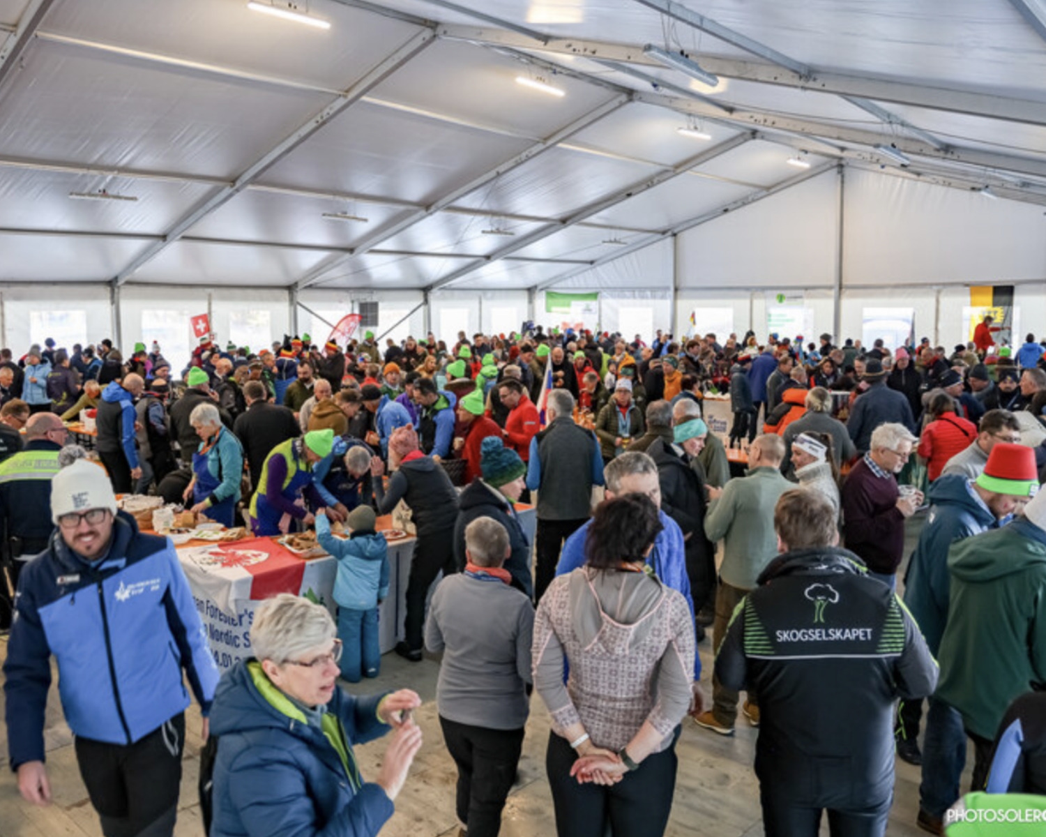 Una grande folla di persone si riunisce all'interno di una tenda spaziosa, socializzando e visitando i tavoli con cibo ed esposizioni. Molti indossano giacche e cappelli e l'atmosfera appare vivace e comunitaria.